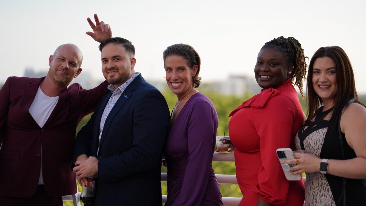 Group of five diverse adults smiling and socializing outdoors, some holding drinks, on a balcony or terrace with blurred cityscape background.