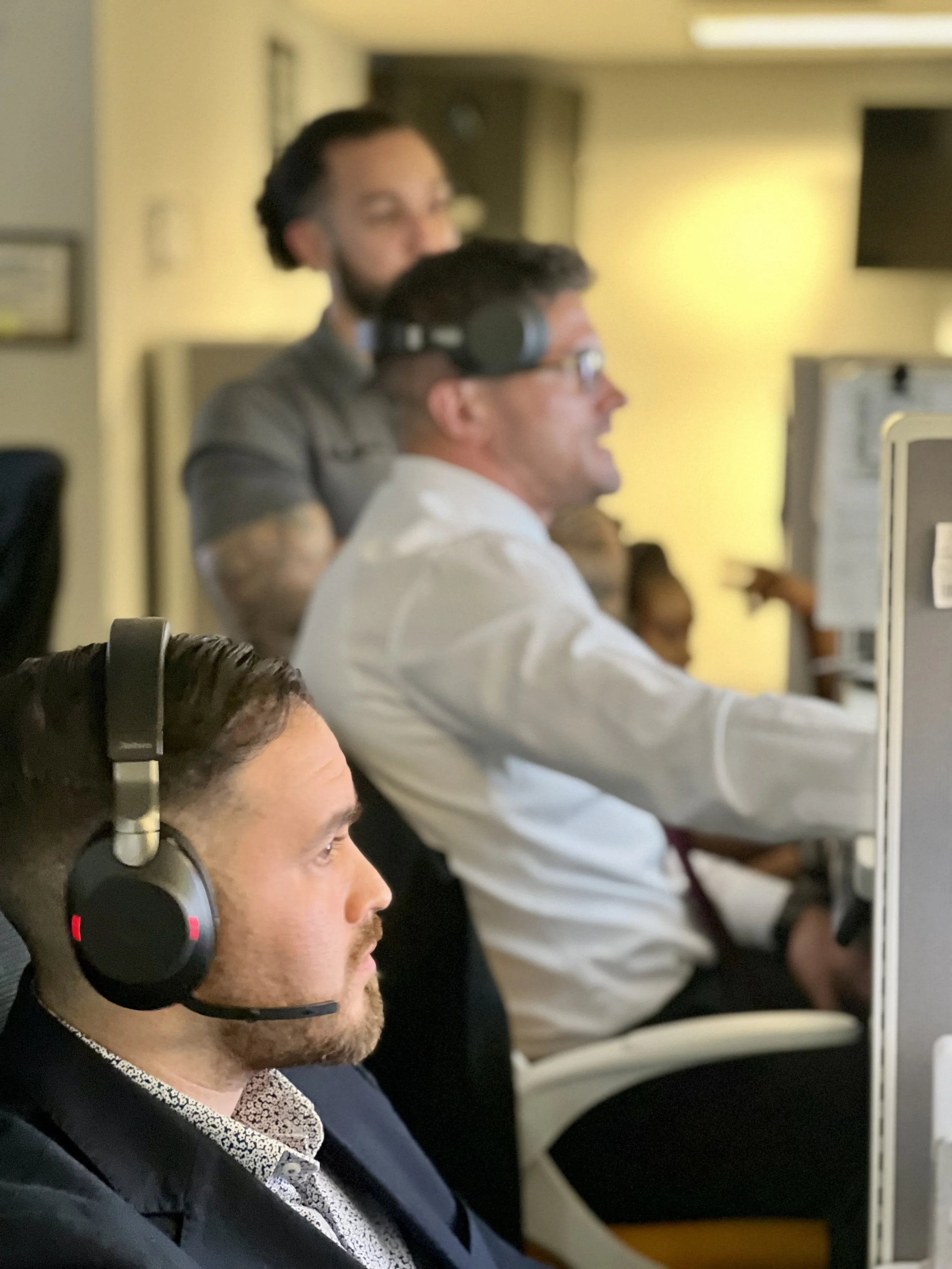 Three professionals working at their desks, wearing headsets, with computer monitors in front of them in a busy office environment.