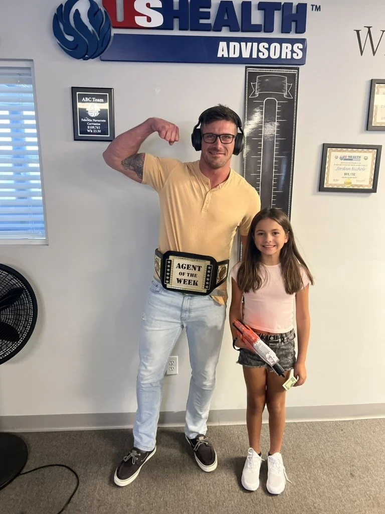 A man and a young girl posing together indoors. The man is flexing his arm and wearing glasses, a yellow t-shirt, jeans, sneakers, and a belt that reads 'Agent of the Week.' The girl is smiling and holding a toy gun. Behind them, there are framed cer