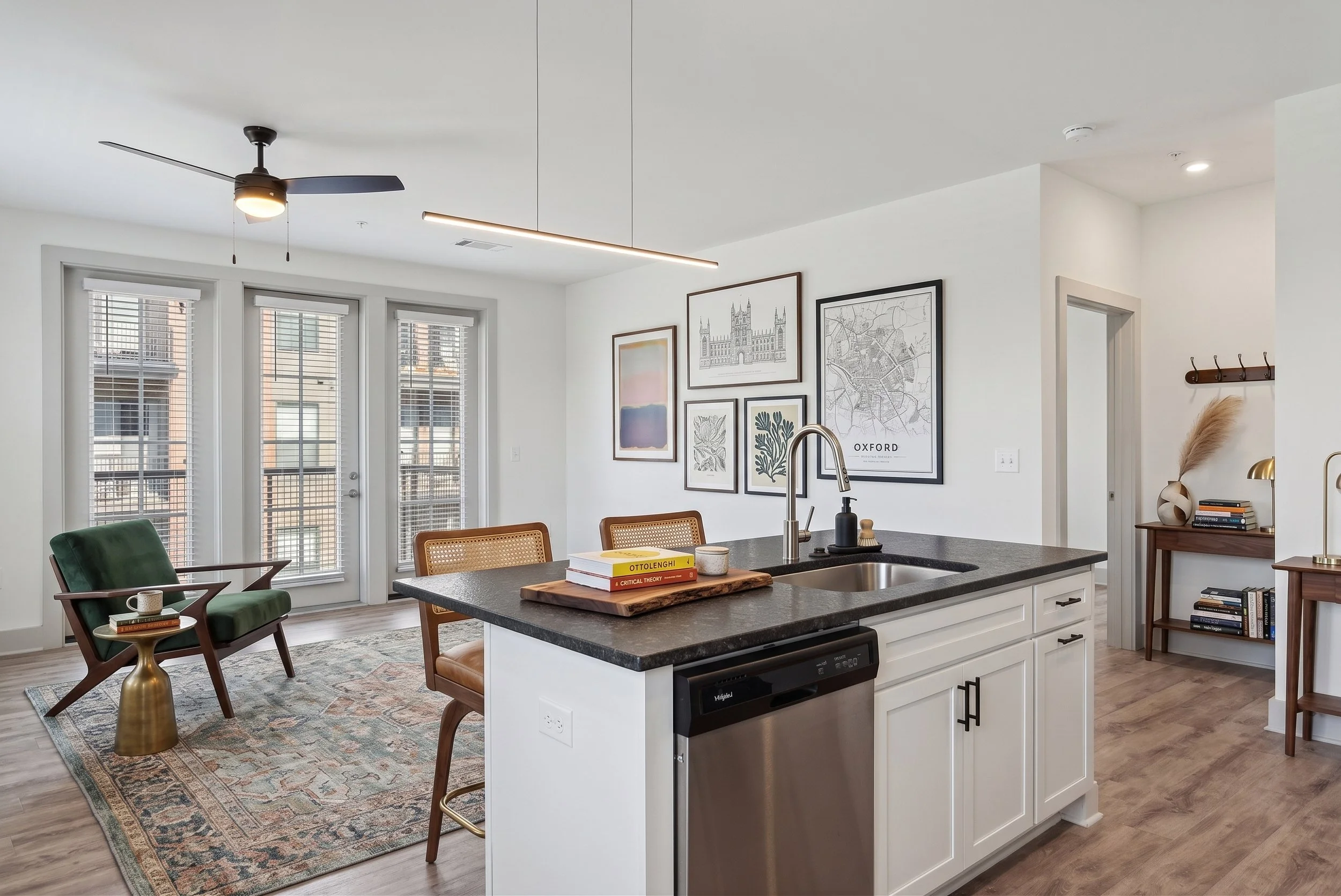 A modern kitchen with a black countertop island, white cabinets, and a stainless steel dishwasher. Behind the island, there's a wall with framed artwork, including a map labeled 'Oxford.' To the left, there are two green armchairs with a small golden