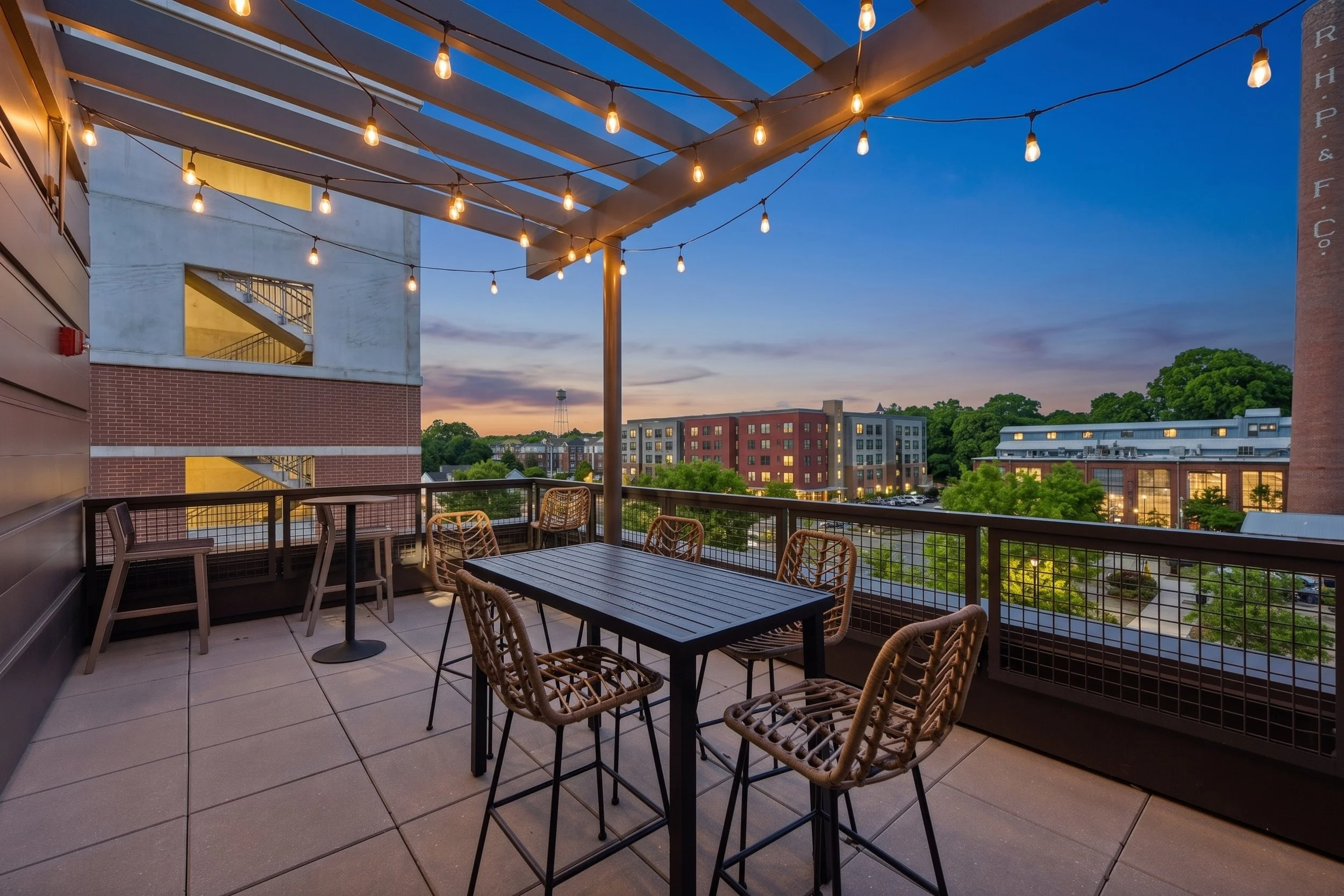 Large outdoor balcony with string lights, a table, and chairs overlooking University Center in Rock Hill, SC at dusk with buildings and trees.