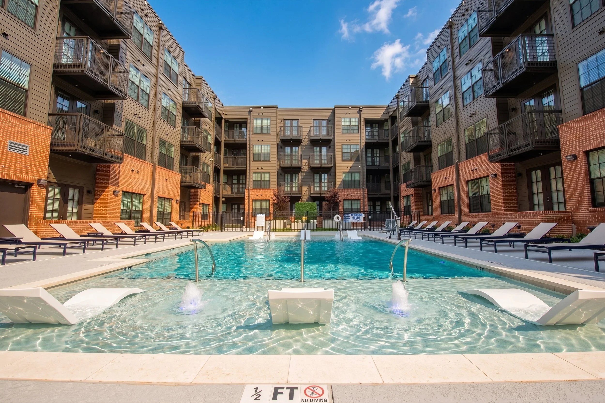 Apartment complex with a swimming pool in the foreground and multi-story residential buildings behind, under a partly cloudy blue sky.