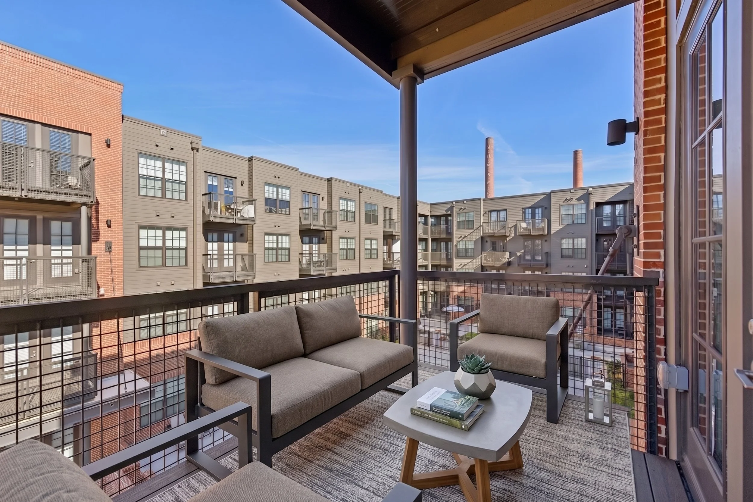 Balcony with outdoor furniture, two beige sofas, a coffee table with books and a small plant, surrounded by metal railing, overlooking a residential apartment complex with multiple-story buildings, brick and gray siding, and tall smokestacks in the background under a clear blue sky.