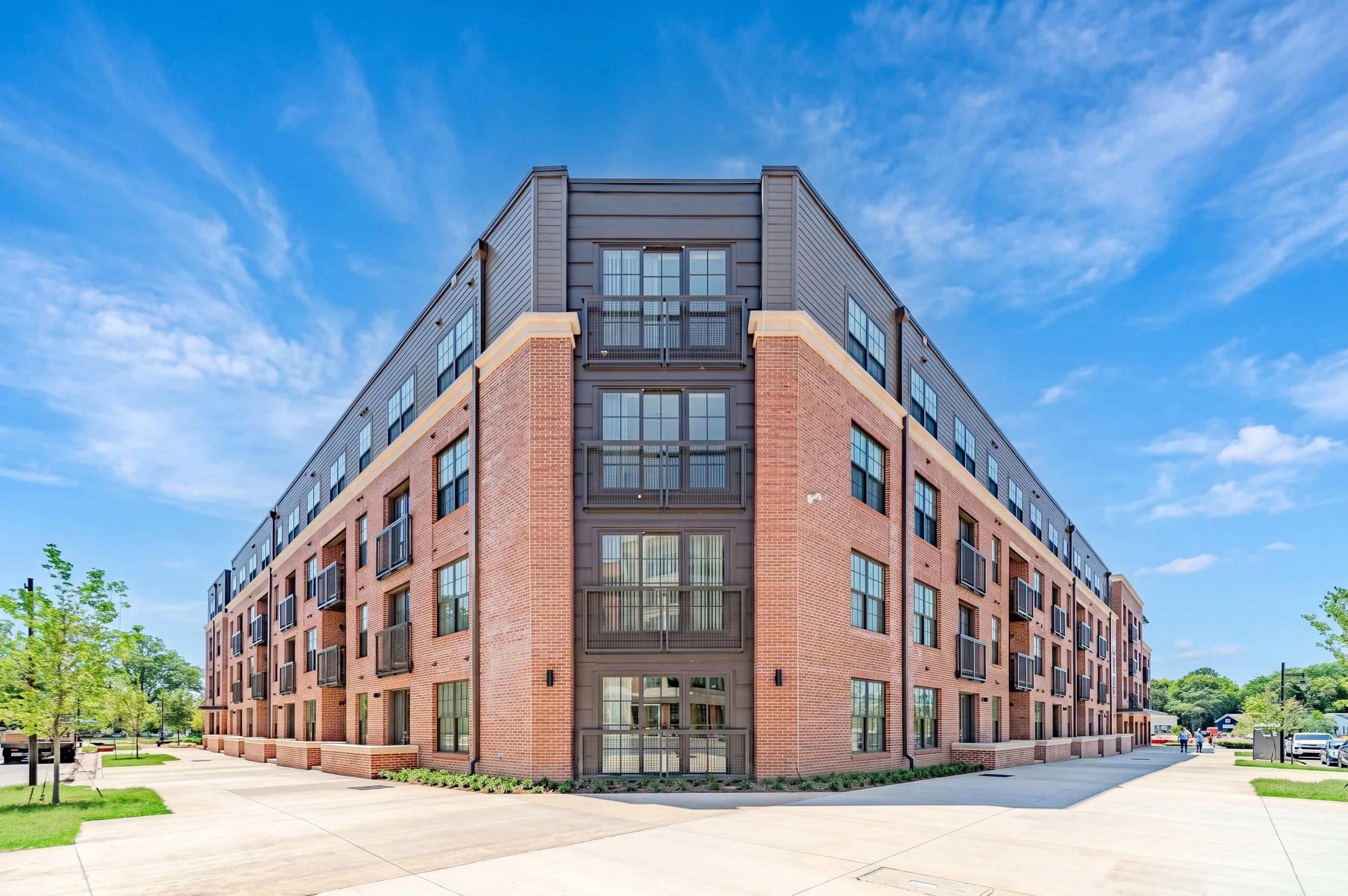 The Easeley is a modern brick apartment building with multiple floors, large windows, and private balconies, surrounded by trees and a sidewalk, under a blue sky with scattered clouds.