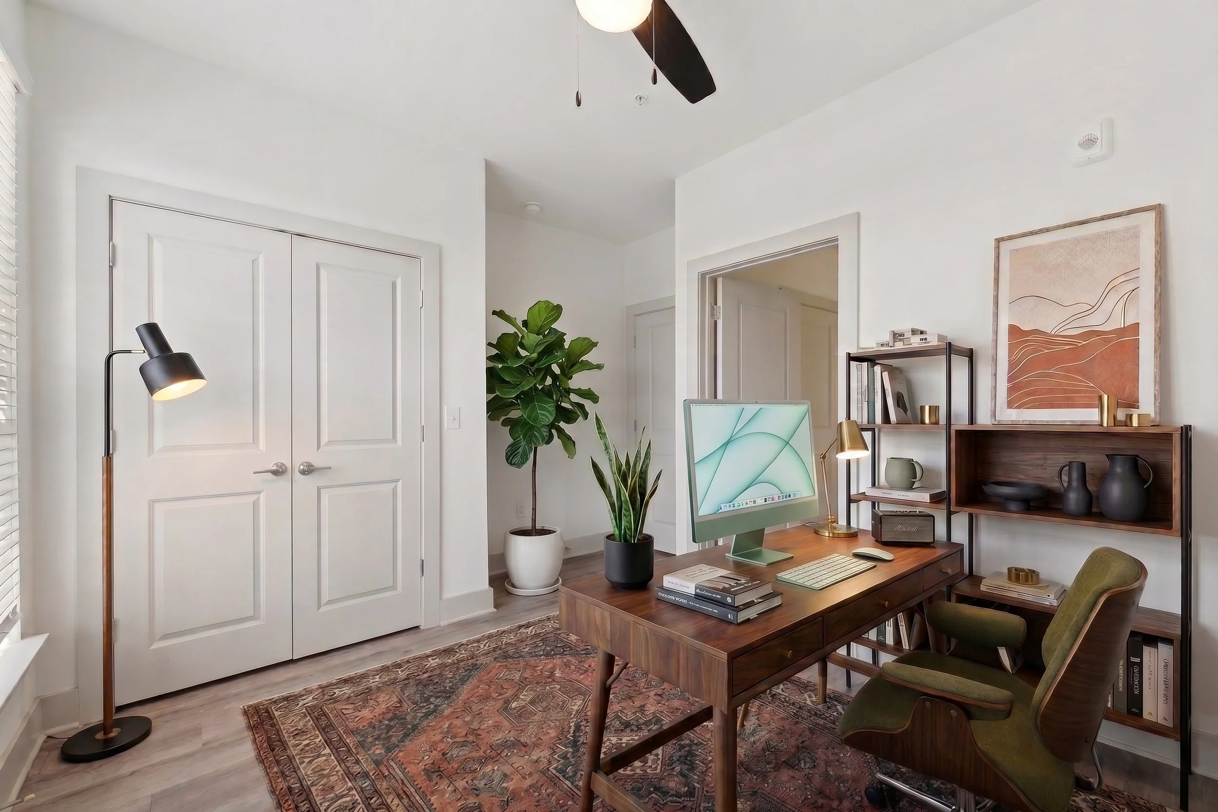 A home office with white walls, a ceiling fan, a tall green plant in a white pot, a wooden desk with an iMac computer, books, and decorative items, a green upholstered chair. There's a black floor lamp, a shelving unit with art, books, and vases, and an ornate area rug.
