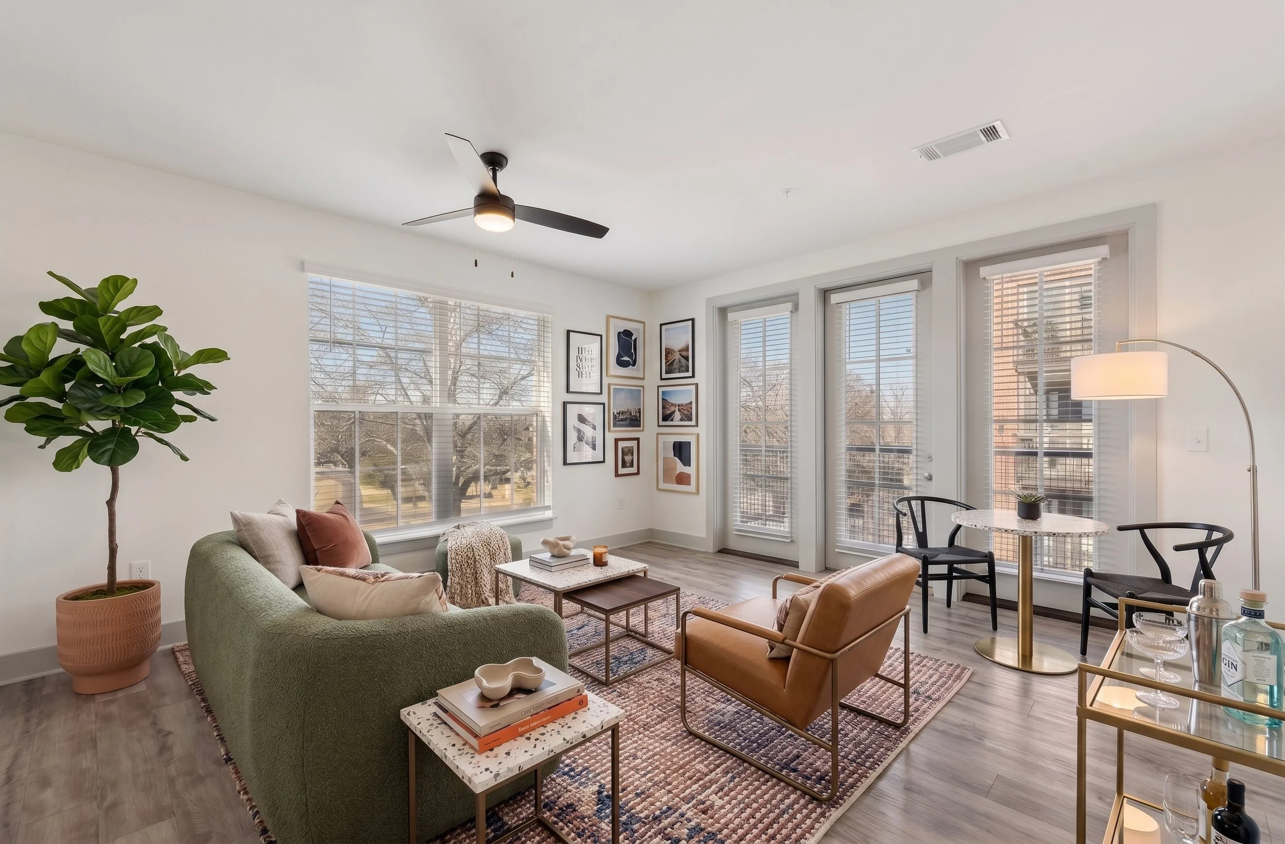 Bright living room with large windows and modern decor, including a green sofa, brown leather armchair, coffee table, and wall art, illuminated by natural sunlight and a floor lamp.