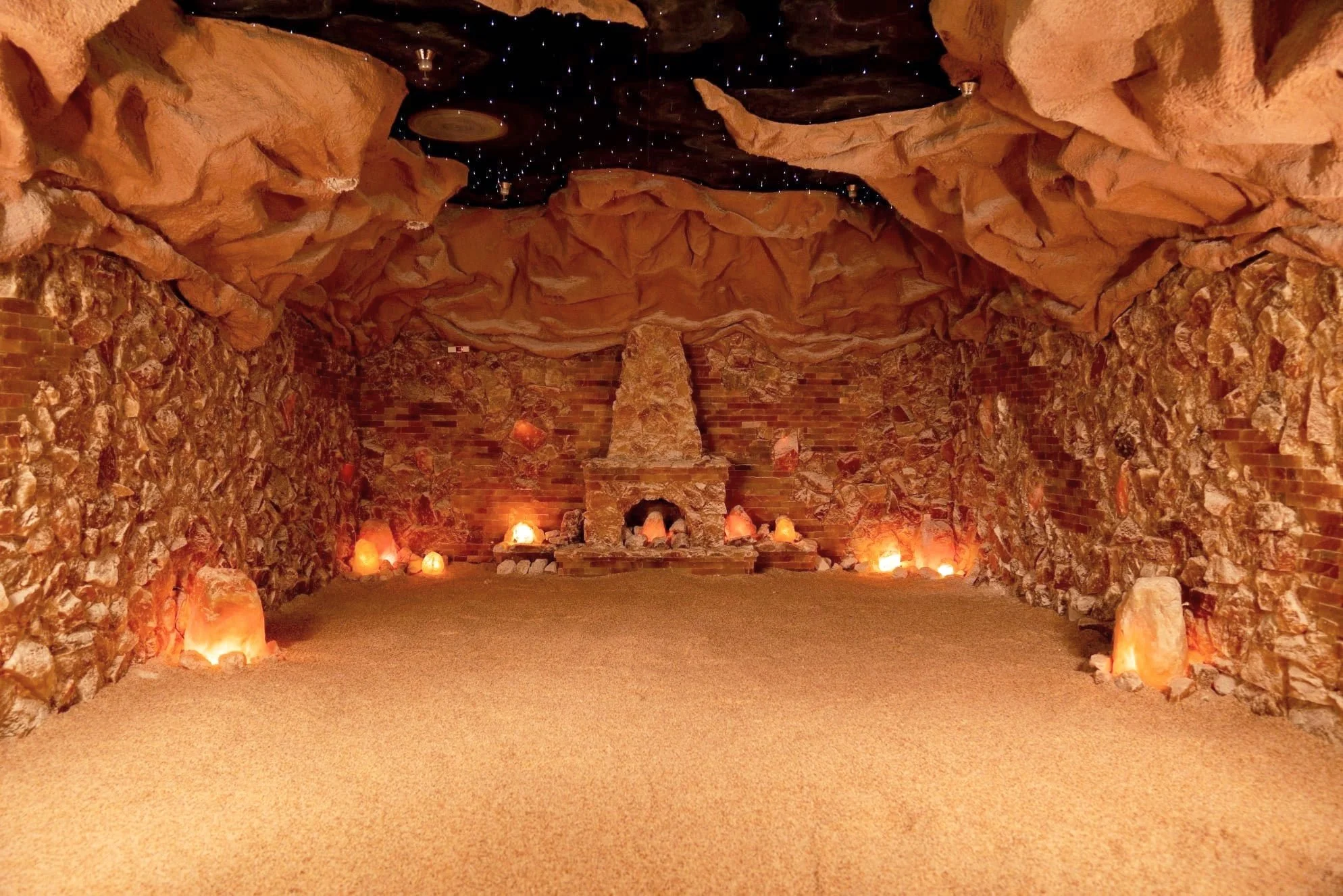 A cozy indoor space designed to resemble a cave, with rock walls and ceiling, a fireplace in the center, and salt lamps lining the floor, creating a warm, soothing glow.