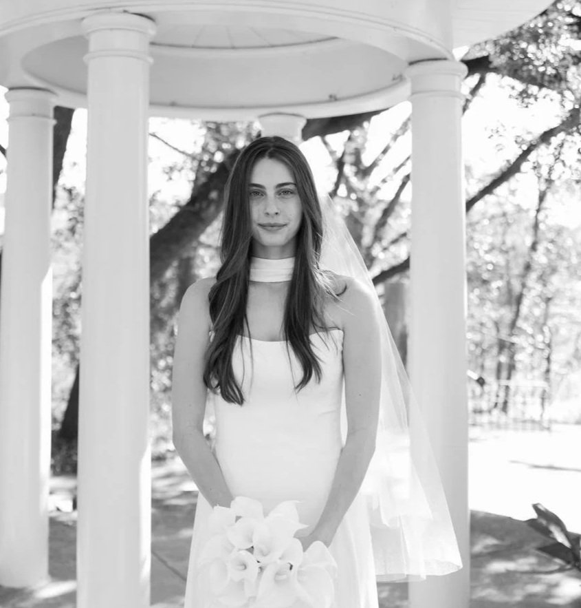 Black and white photo of a young woman in a wedding dress holding a bouquet, standing outdoors near a white gazebo with trees in the background.