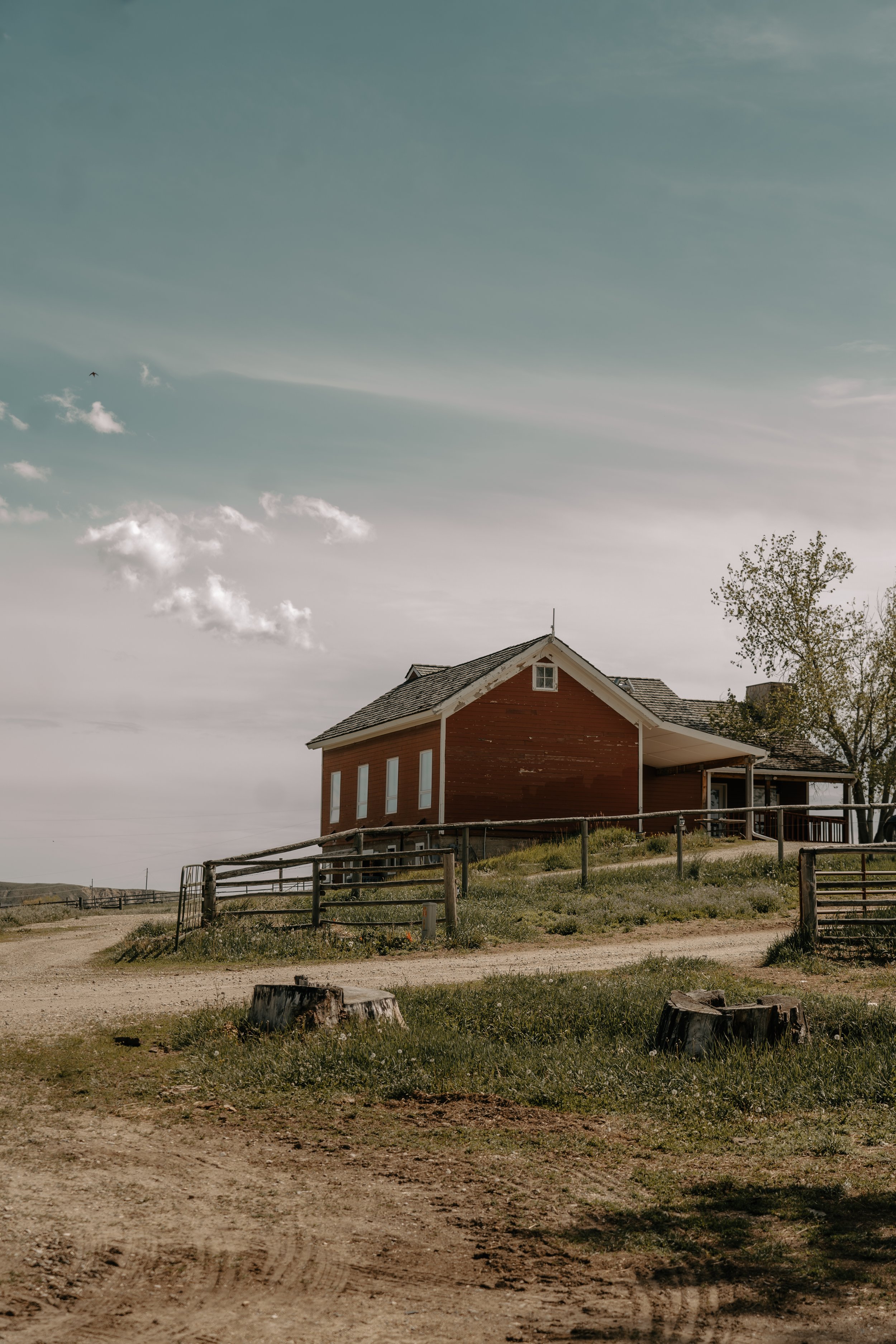 A rural red house on a hillside with a wooden fence, dirt roads, and a tree, under a blue sky with some clouds. TA Ranch, guest ranch, dude ranch, working cattle ranch, horse ranch, horseback riding, Buffalo Wyoming North-eastern Wyoming, homestead,