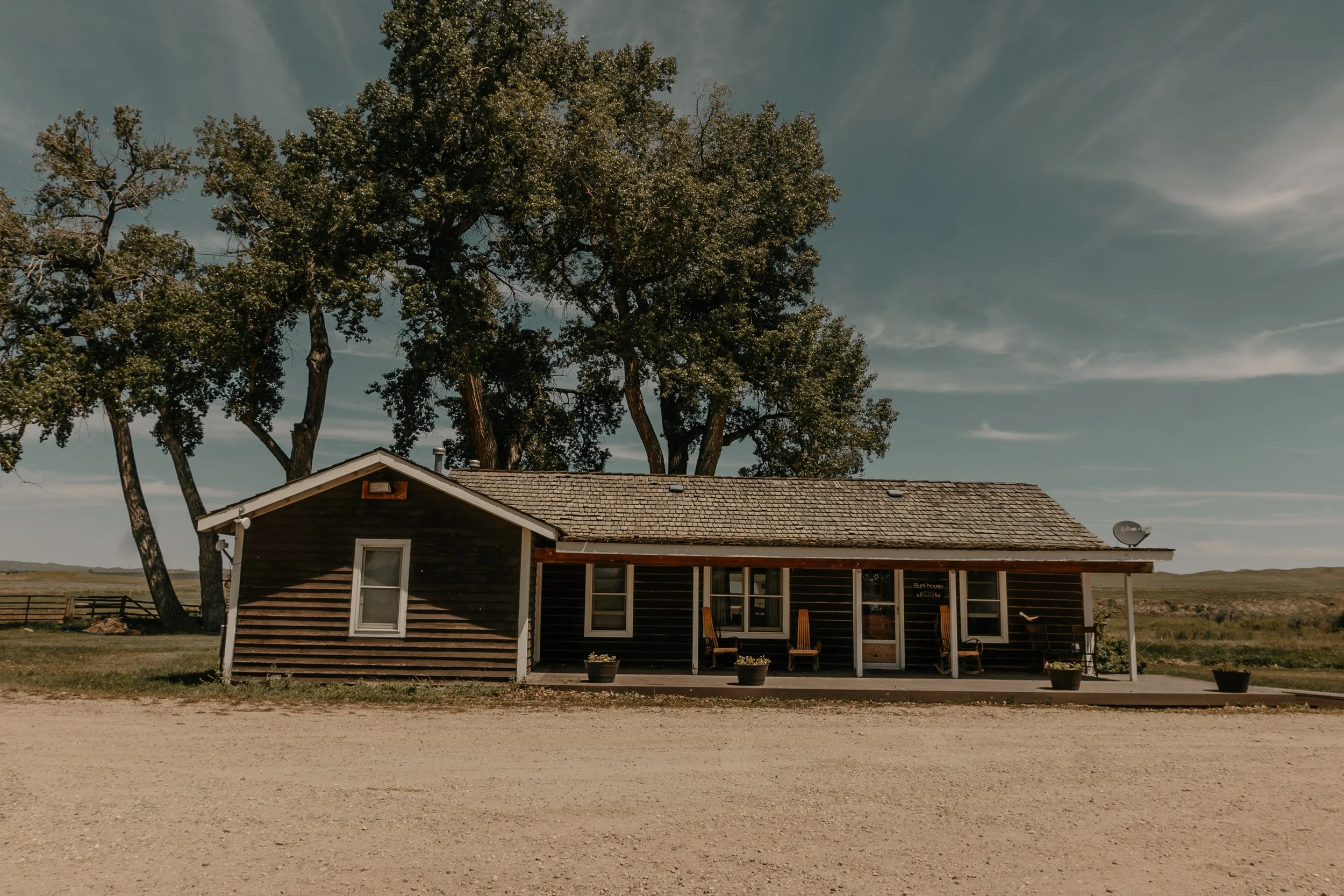 A rustic wooden house with a porch, surrounded by a few potted plants, underneath a large tree with a clear sky overhead in a rural landscape. TA Ranch, guest ranch, dude ranch, working cattle ranch, horse ranch, horseback riding, Buffalo Wyoming