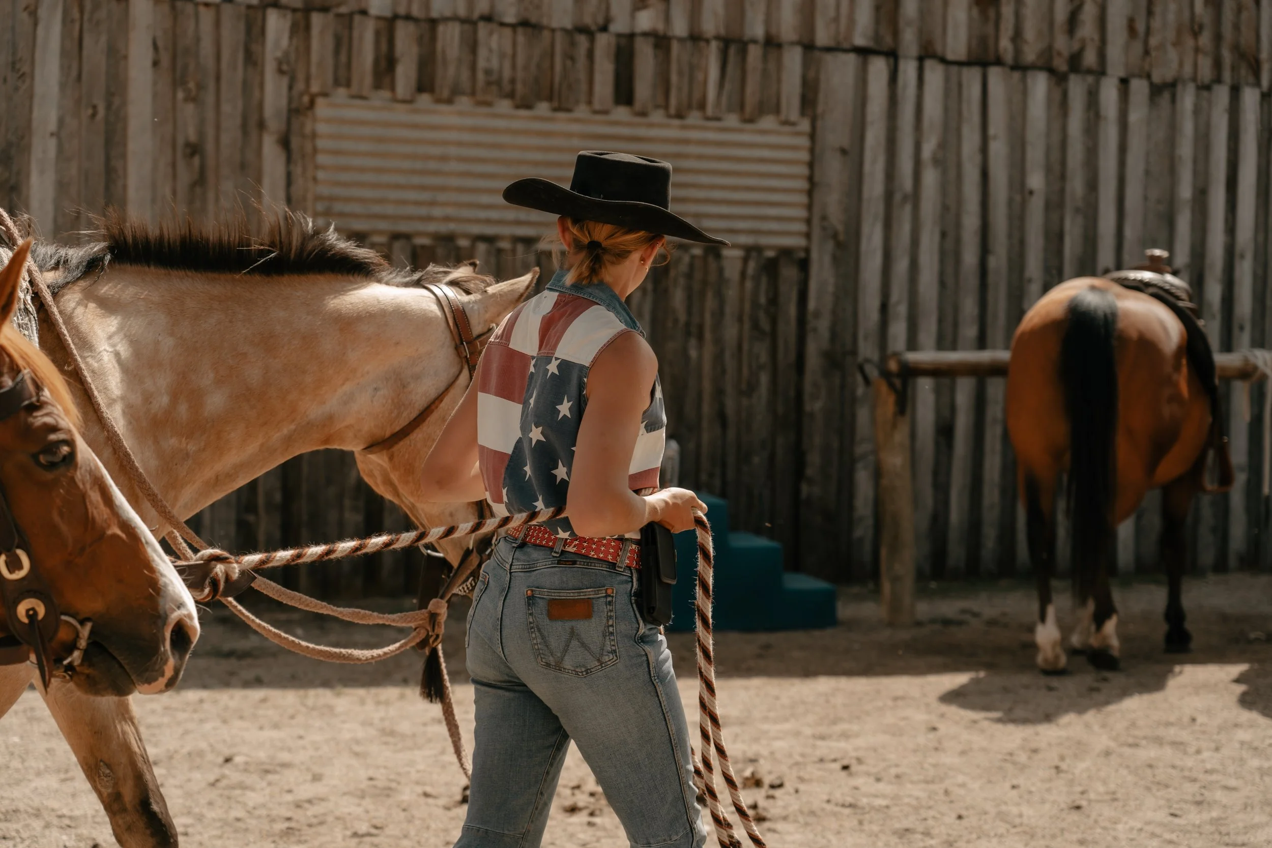 A young woman with a cowboy hat and American flag sleeveless shirt walking a horse in a rustic outdoor corral.