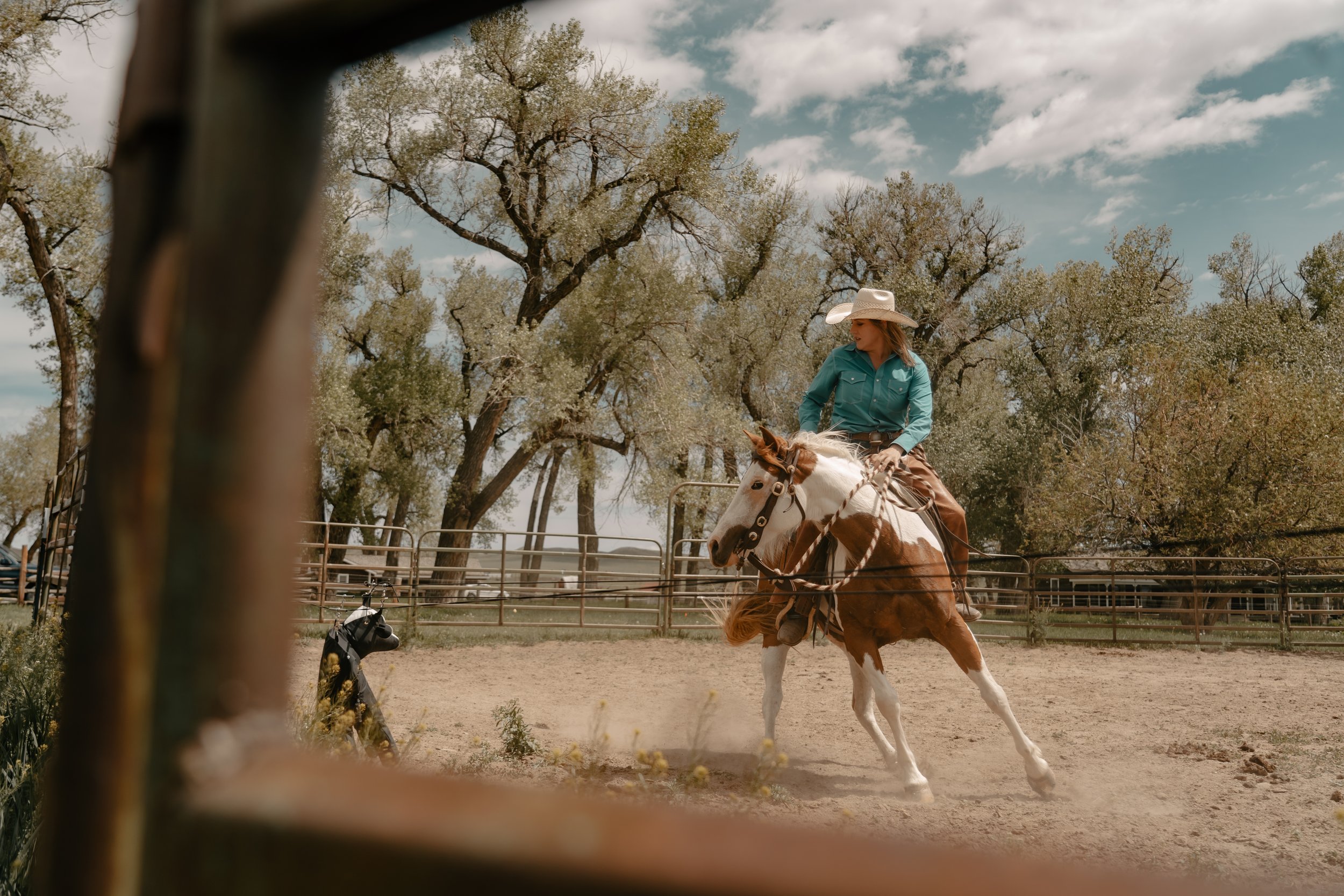A woman riding a pinto horse in an outdoor riding arena, with trees and a fence in the background, clouds in the sky, and a black object in the foreground. TA Ranch, guest ranch, dude ranch, working cattle ranch, horse ranch, horseback riding, WY