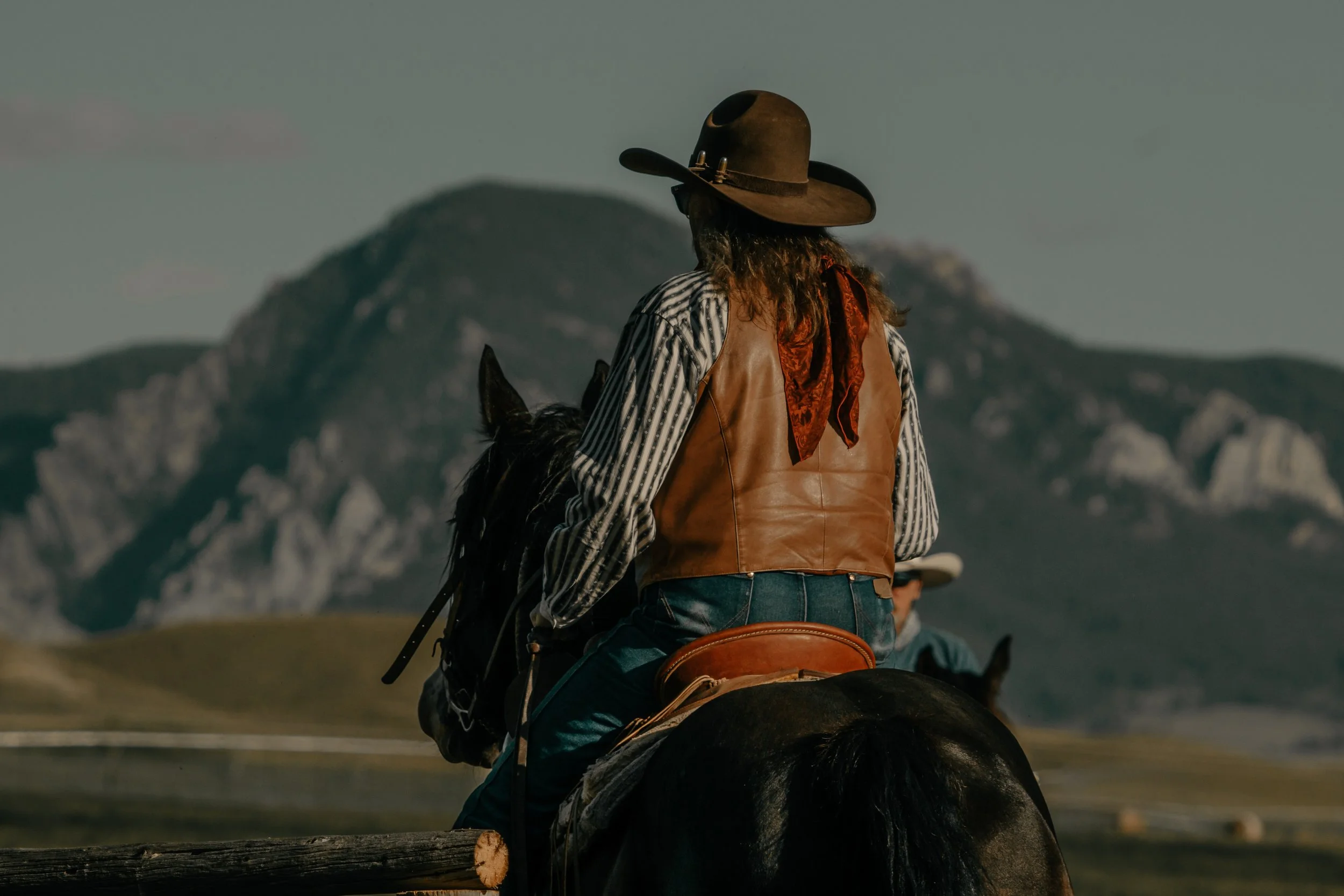 A cowboy wearing a wide-brimmed hat, leather vest, and striped shirt riding a black horse with a mountainous landscape in the background.