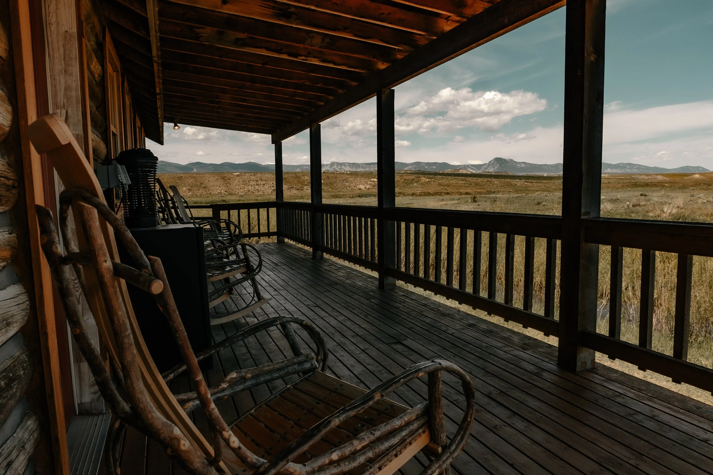 A rustic covered porch with wooden rocking chairs overlooking an expansive open landscape with mountains in the distance under a partly cloudy sky. TA Ranch, guest ranch, dude ranch, working cattle ranch, horse ranch, horseback riding, Buffalo WY