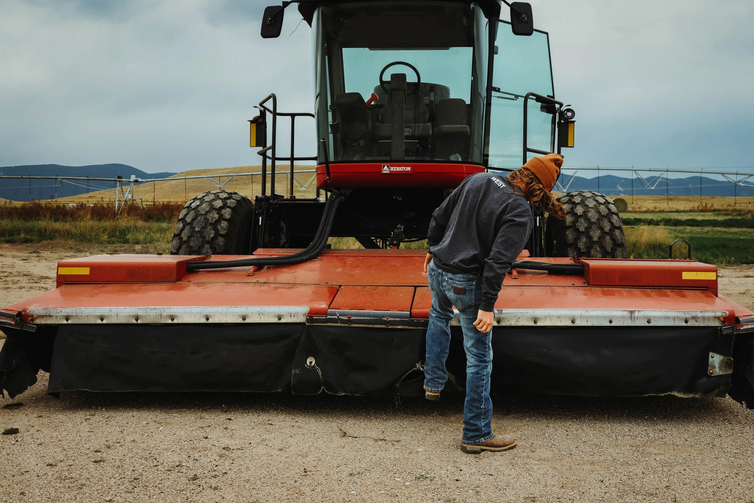 A person inspecting or repairing a large red tractor on a farm field during the day, with irrigation equipment and mountains in the background. TA Ranch, guest ranch, dude ranch, working cattle ranch, horse ranch, horseback riding, Buffalo Wyoming