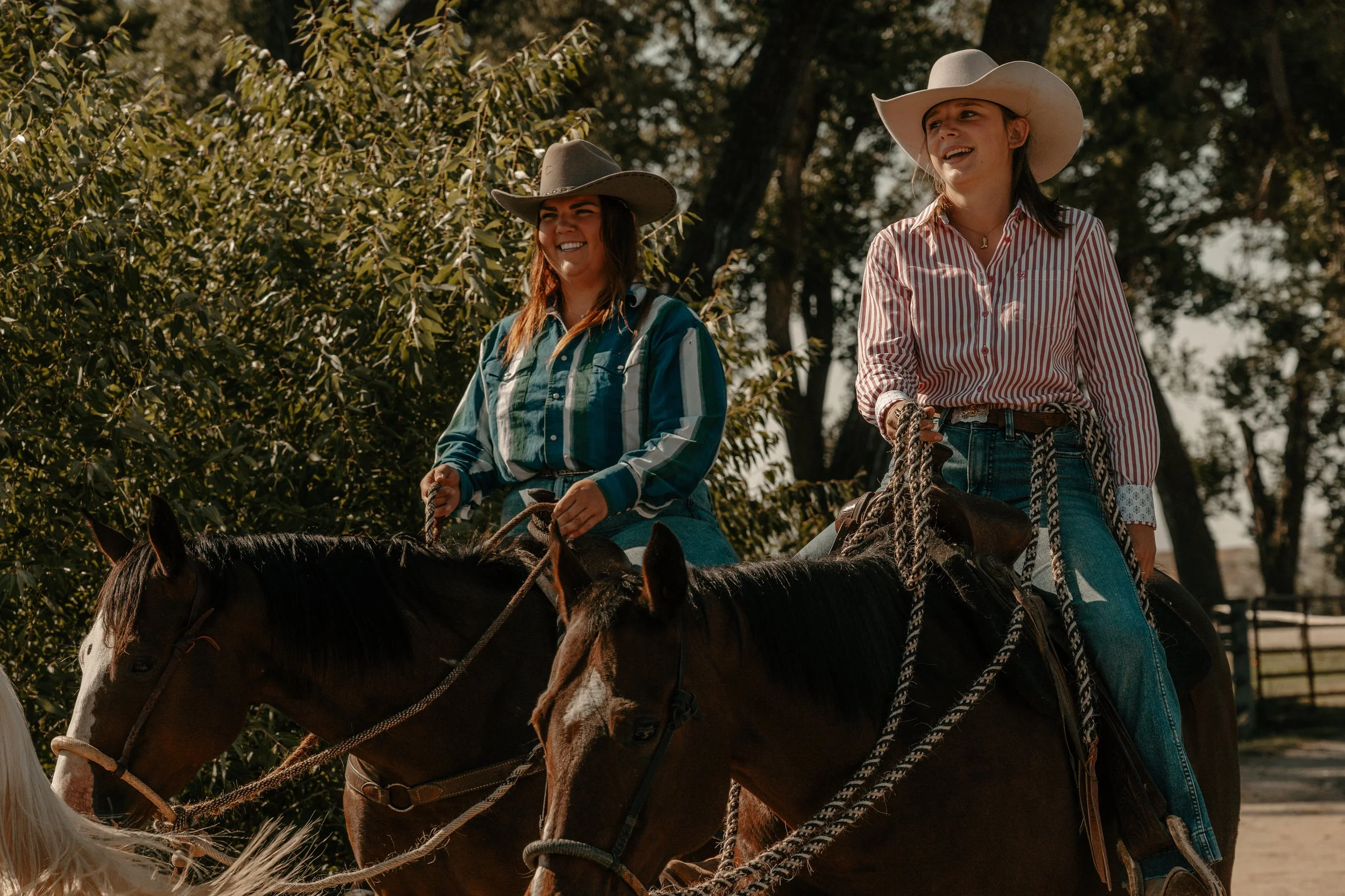 Two women riding horses outdoors on a sunny day, both wearing cowboy hats and casual button-up shirts, with trees in the background. TA Ranch, guest ranch, dude ranch, working cattle ranch, horse ranch, horseback riding, Buffalo Wyoming, agriculture