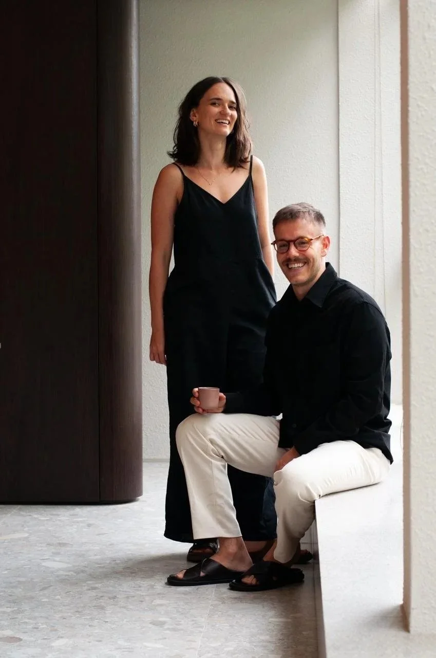 Um homem sentado em uma banqueta com um copo na mão, ao seu lado uma mulher de vestido preto sorrindo, ambos posando para a câmera em um ambiente interno.