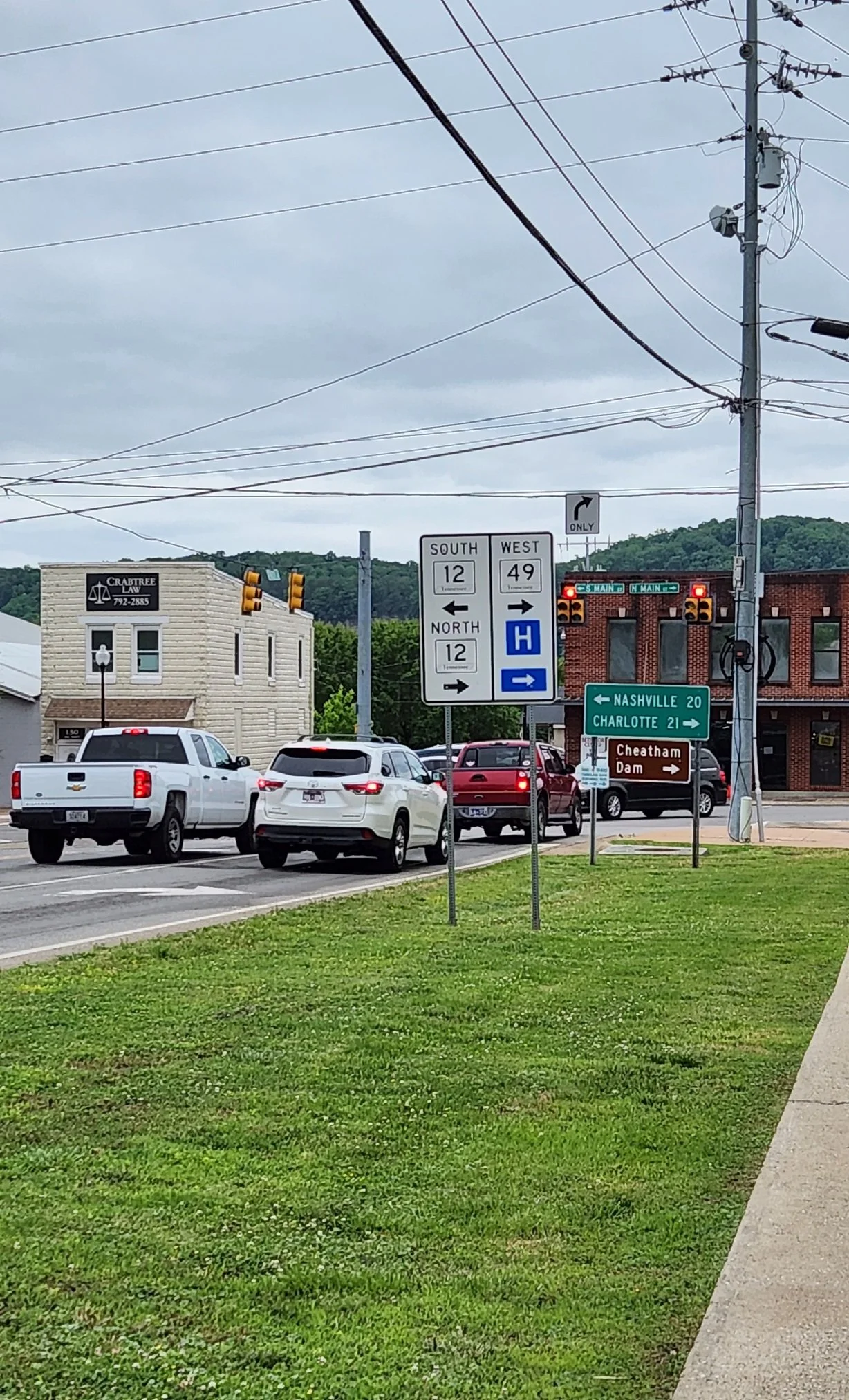 Traffic intersection with directional signs for highways 12 and 49, pointing towards Nashville, Charlotte, and Cheatham Dam, along with a hospital direction sign. Cars are waiting at a traffic light near buildings.