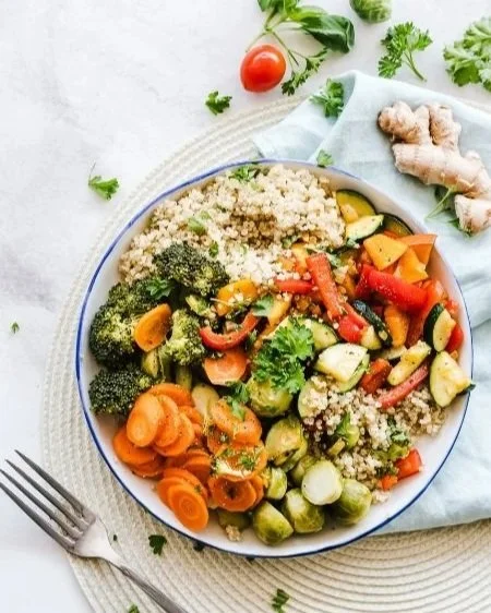 A bowl of mixed vegetables, quinoa, and carrots on a white plate. Fresh vegetables, ginger, cherry tomato, and parsley are visible on the table around the plate.