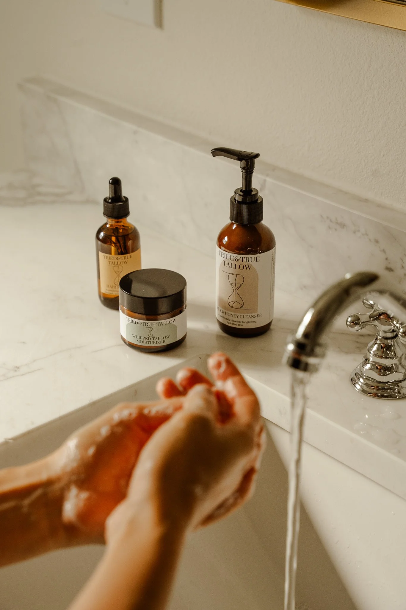Person washing hands at bathroom sink with skincare products nearby, including a honey cleanser, moisturizer, and serum on the countertop.