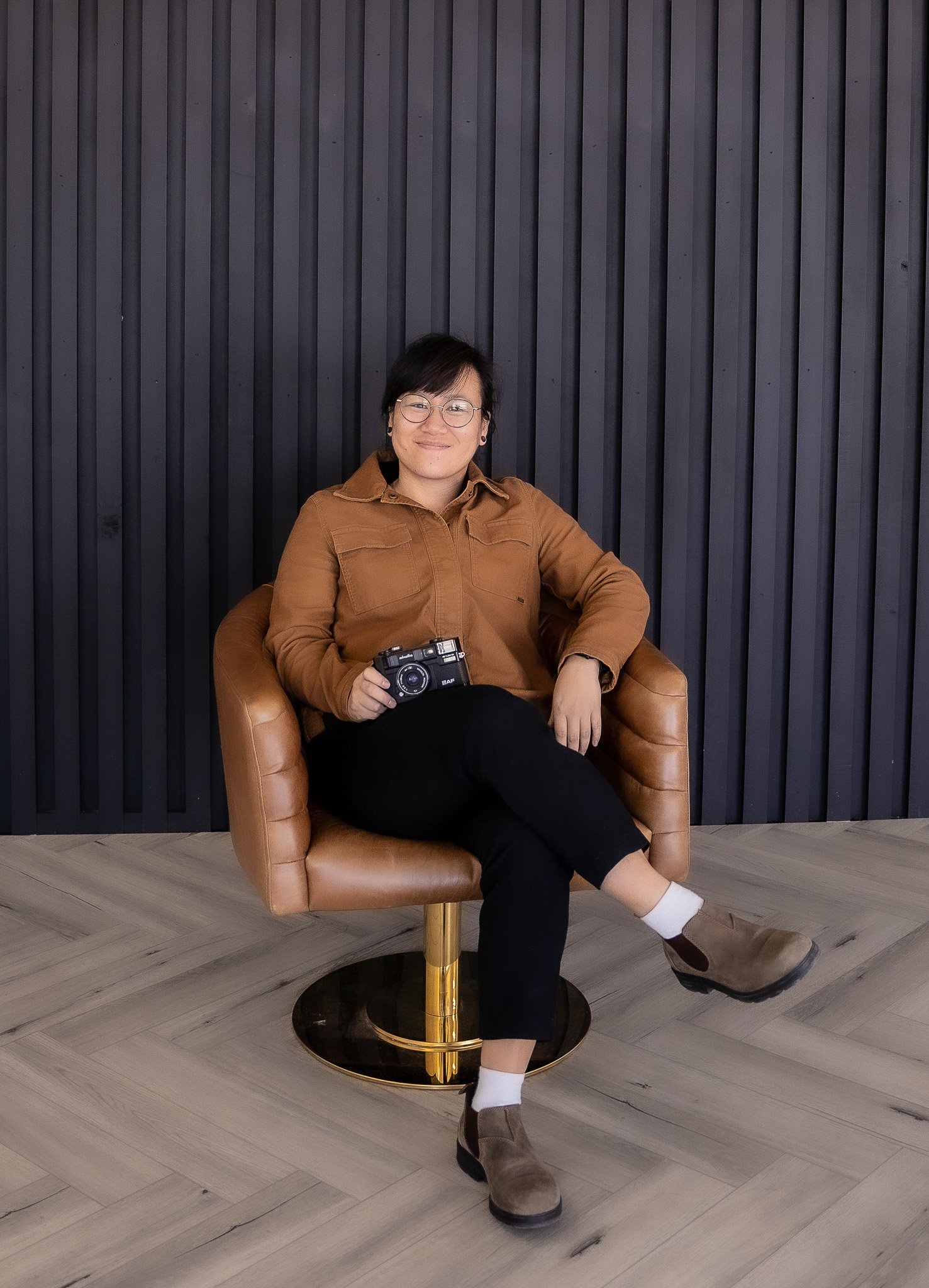 A woman sitting in a brown leather armchair against a dark vertical panel wall, holding a vintage camera, dressed in a brown jacket, black pants, brown boots, and white socks, smiling.
