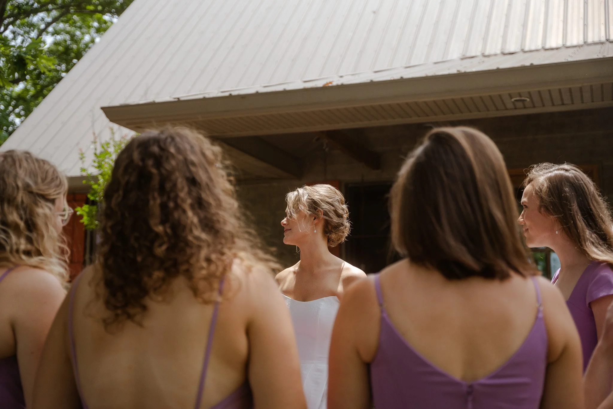 A group of women in purple dresses standing outdoors in front of a wooden building, with one woman in the center wearing a white dress.