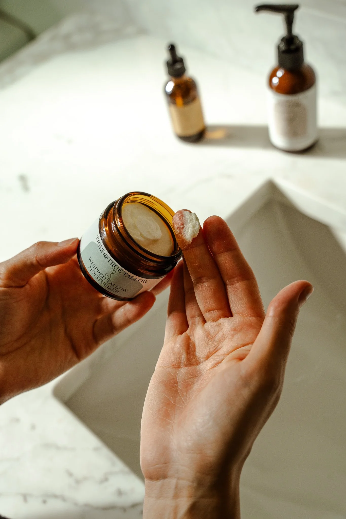 Person holding a jar of moisturizer and applying a small amount of product to their finger. Two additional bottles of skincare products are on the counter in the background.