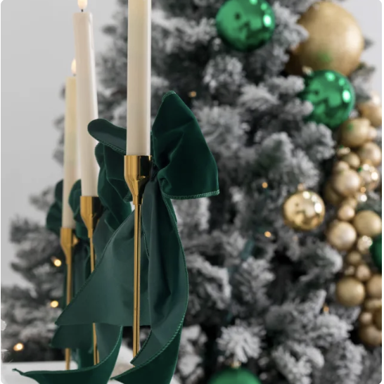 Close-up of gold candlestick holders with white candles and green ribbons, with a decorated Christmas tree in the background.