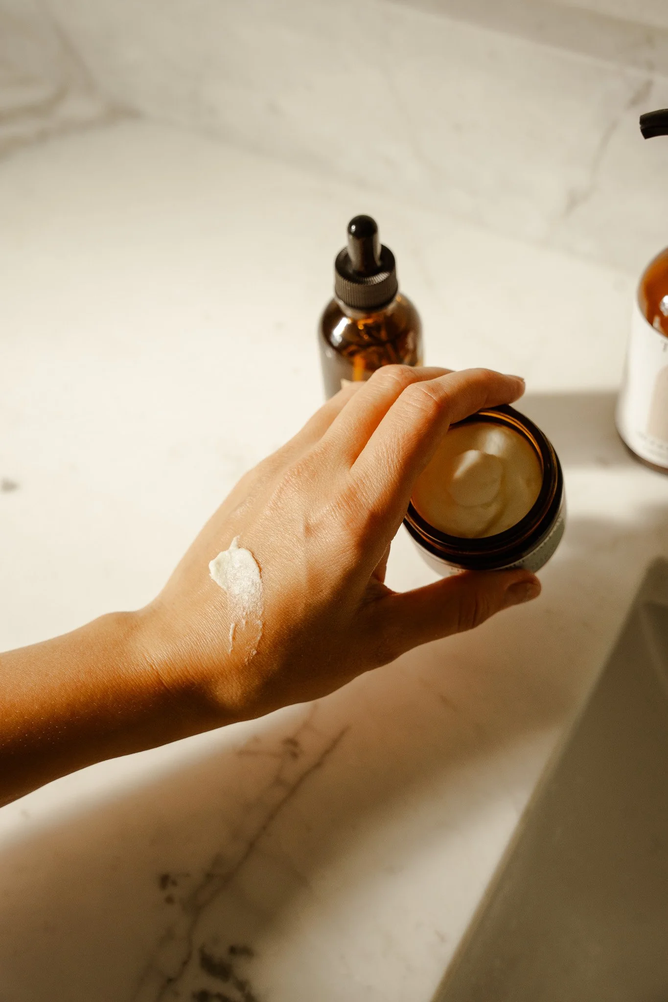 A person with light skin applying cream from an open jar onto the back of their hand. In the background, there are two brown bottles, one with a dropper cap, on a white marble surface.