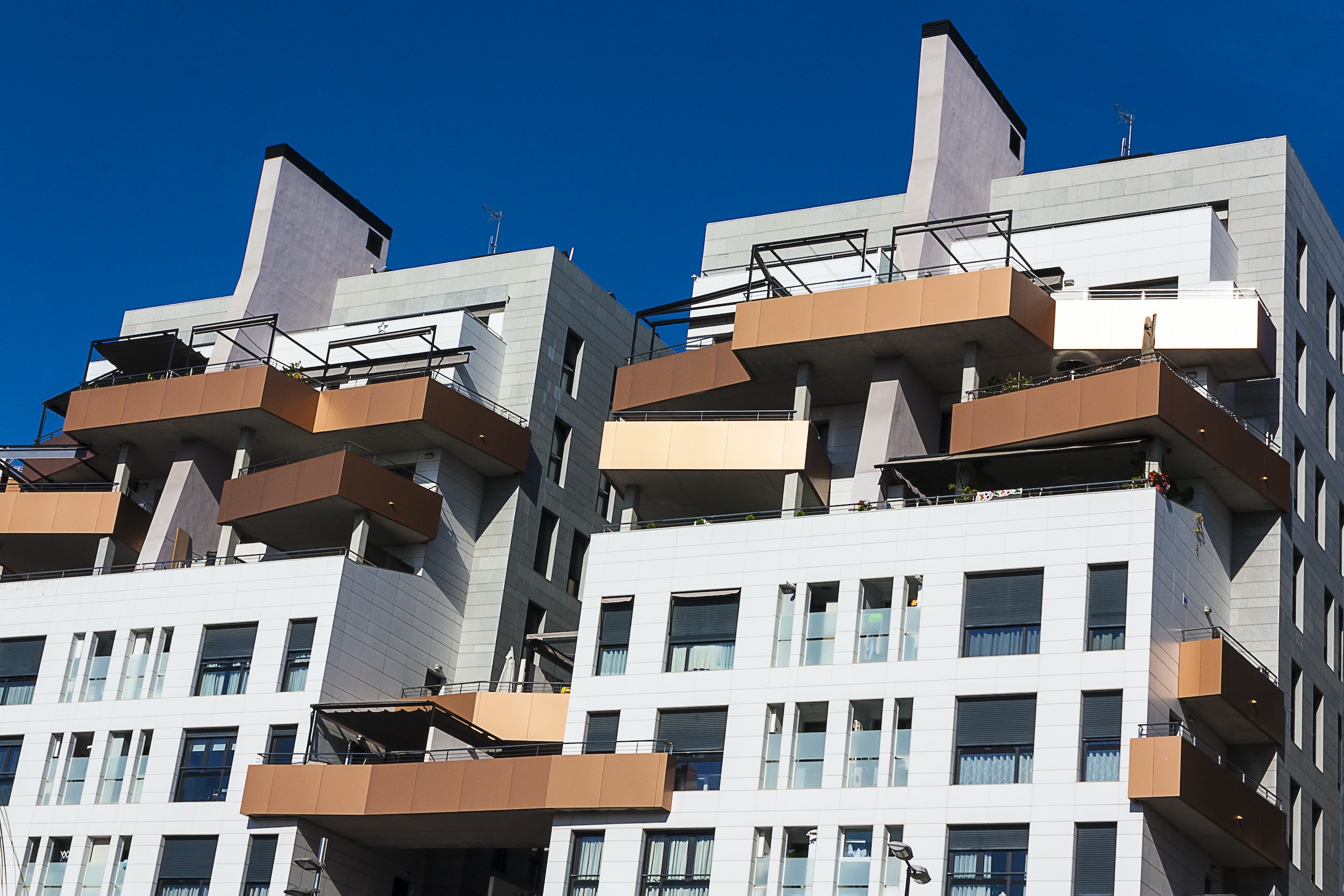 Modern multi-story apartment building with white and gray exterior, featuring balcony areas with orange accents and glass railings, set against a clear blue sky.
