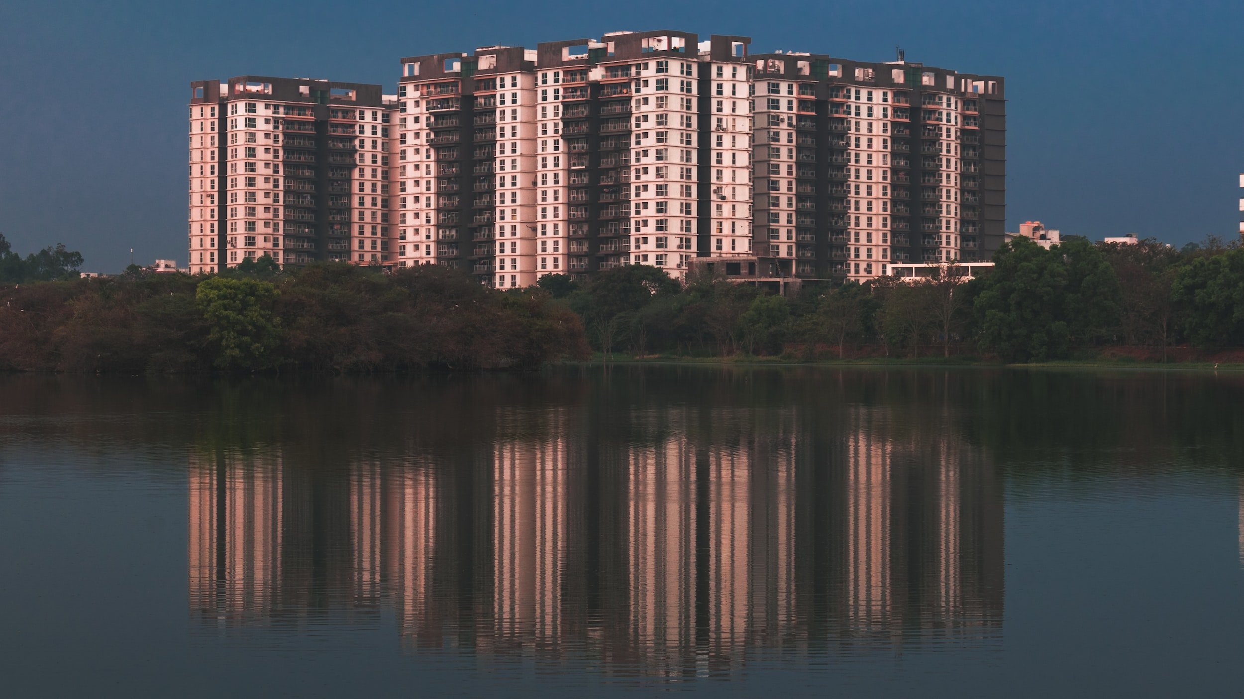 A group of high-rise apartment buildings reflected in a body of water with trees in the foreground.