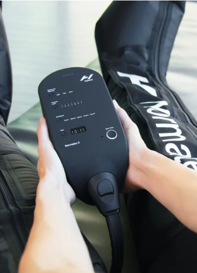 Person holding a black tennis ball machine with digital display and control buttons, next to a tennis racket bag.