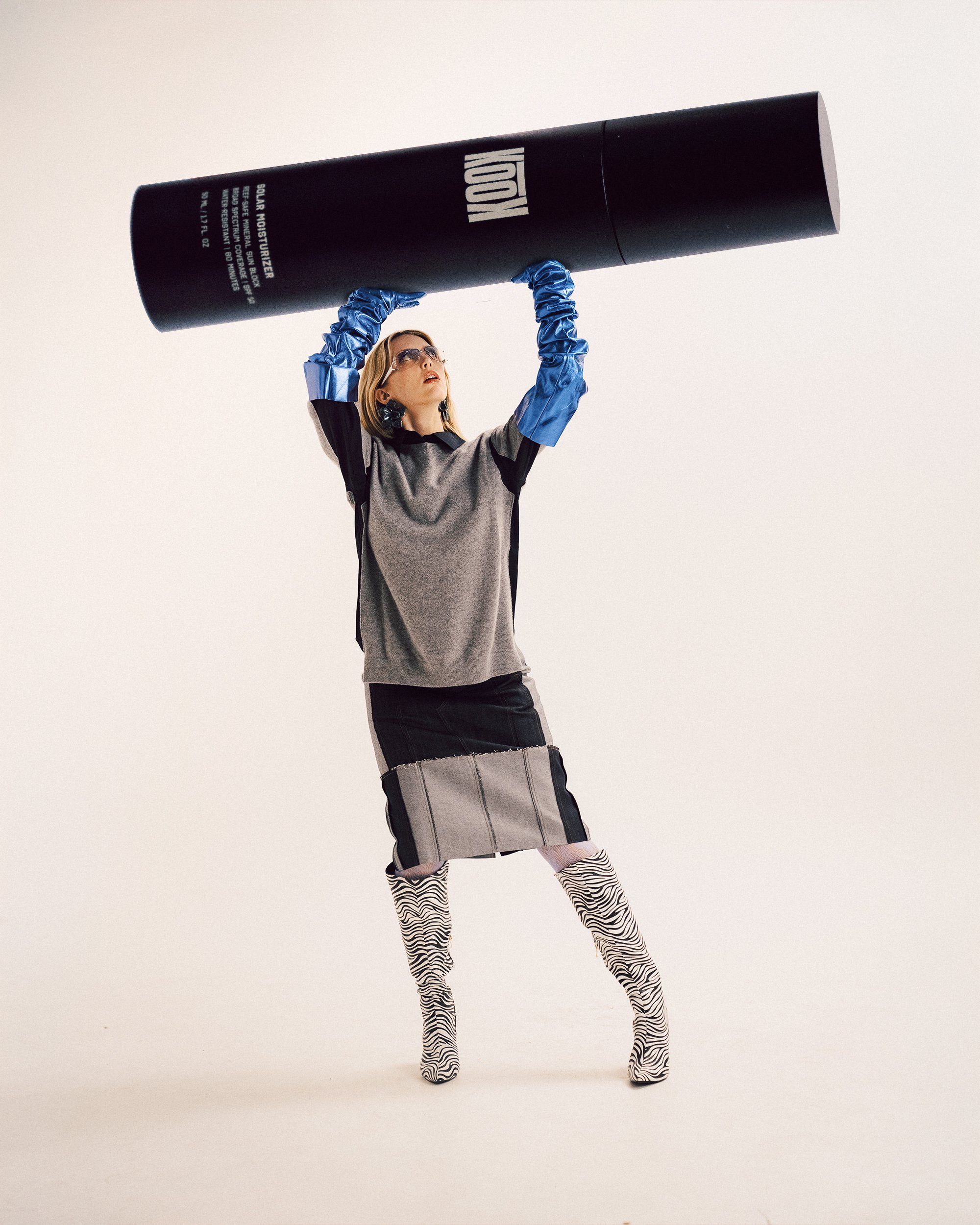 A blonde woman holding up a giant black 'Krave Beauty' product bottle, wearing a grey and dark outfit with blue gloves, glasses, and zebra-print boots.