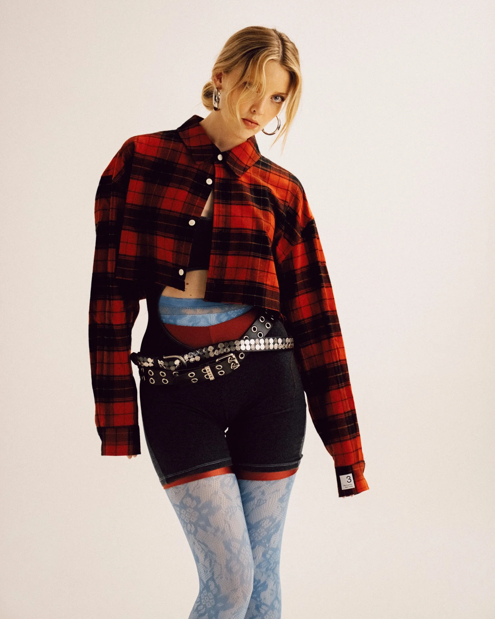 A blonde woman standing and looking down towards the camera, wearing a red and black plaid cropped button-down shirt, dark shorts over patterned tights, double belts, and hoop earrings