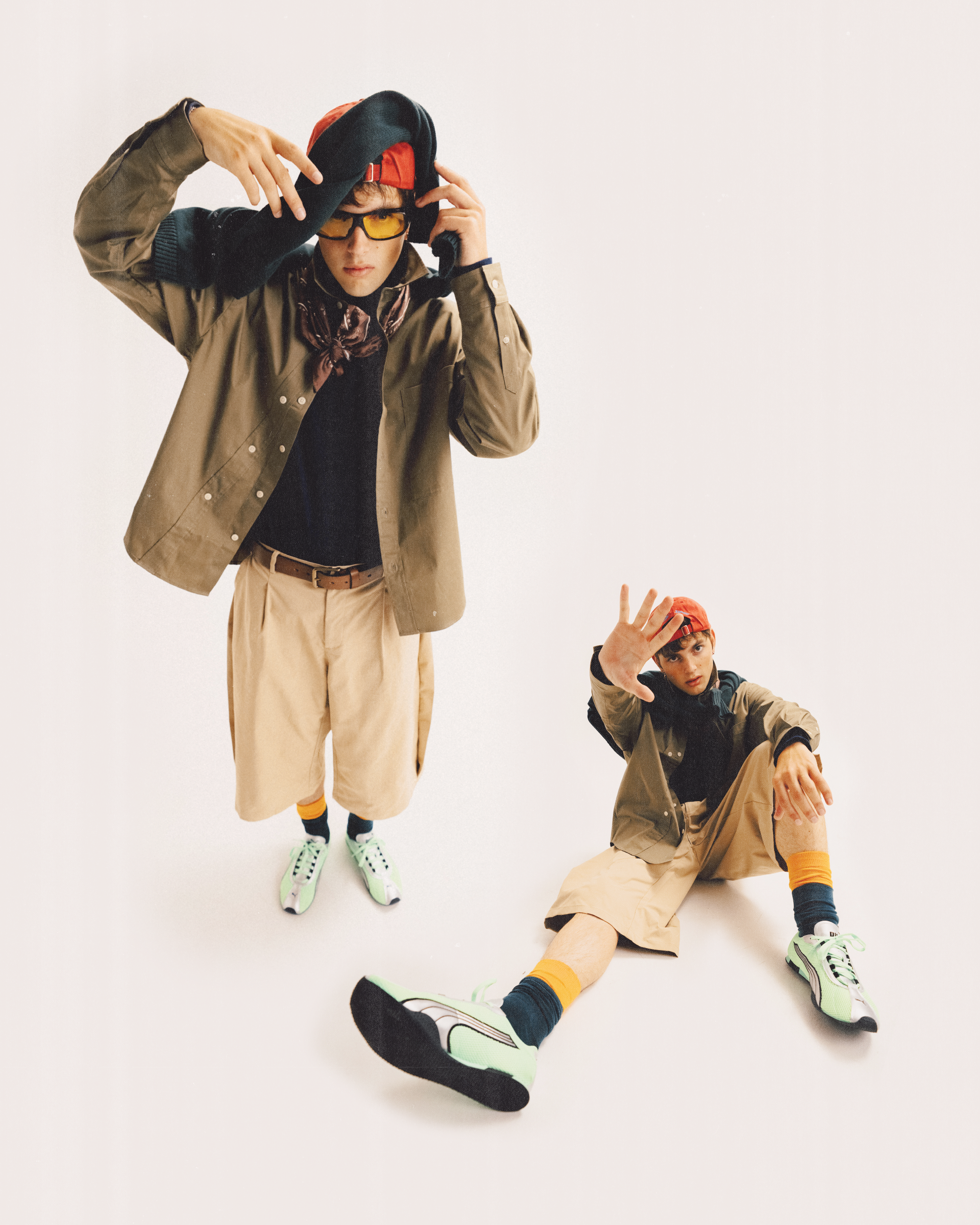 A composite portrait of a young man in two poses against a white background, wearing a dark top, an unbuttoned jacket, khaki shorts, a belt, patterned socks, light-colored sneakers, tinted glasses, a neck scarf, and a red cap.