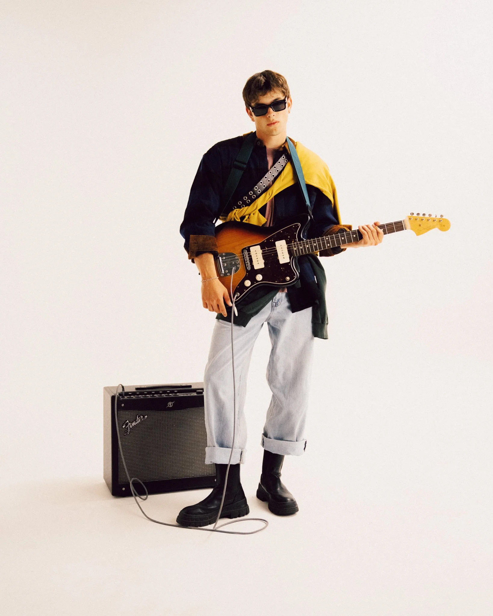 A young man standing and holding an electric guitar next to an amplifier, wearing a dark patterned shirt with a contrasting collar, a yellow garment draped over his shoulder, light-colored cuffed jeans, dark boots, and sunglasses.