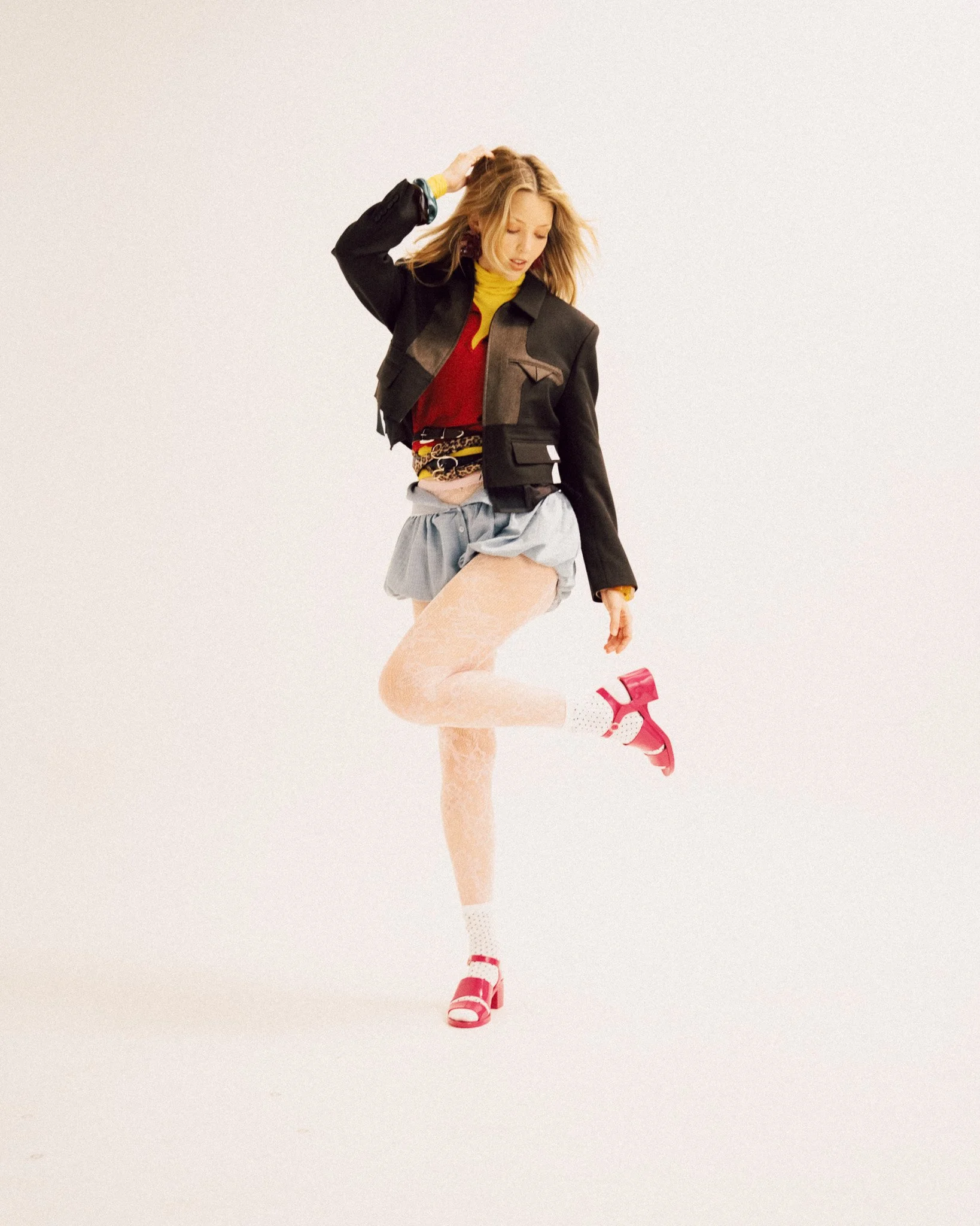 A blonde woman standing on one leg while lifting the other foot, wearing a black jacket over layered tops, light-colored ruffled shorts, patterned tights, white socks, and pink sandals