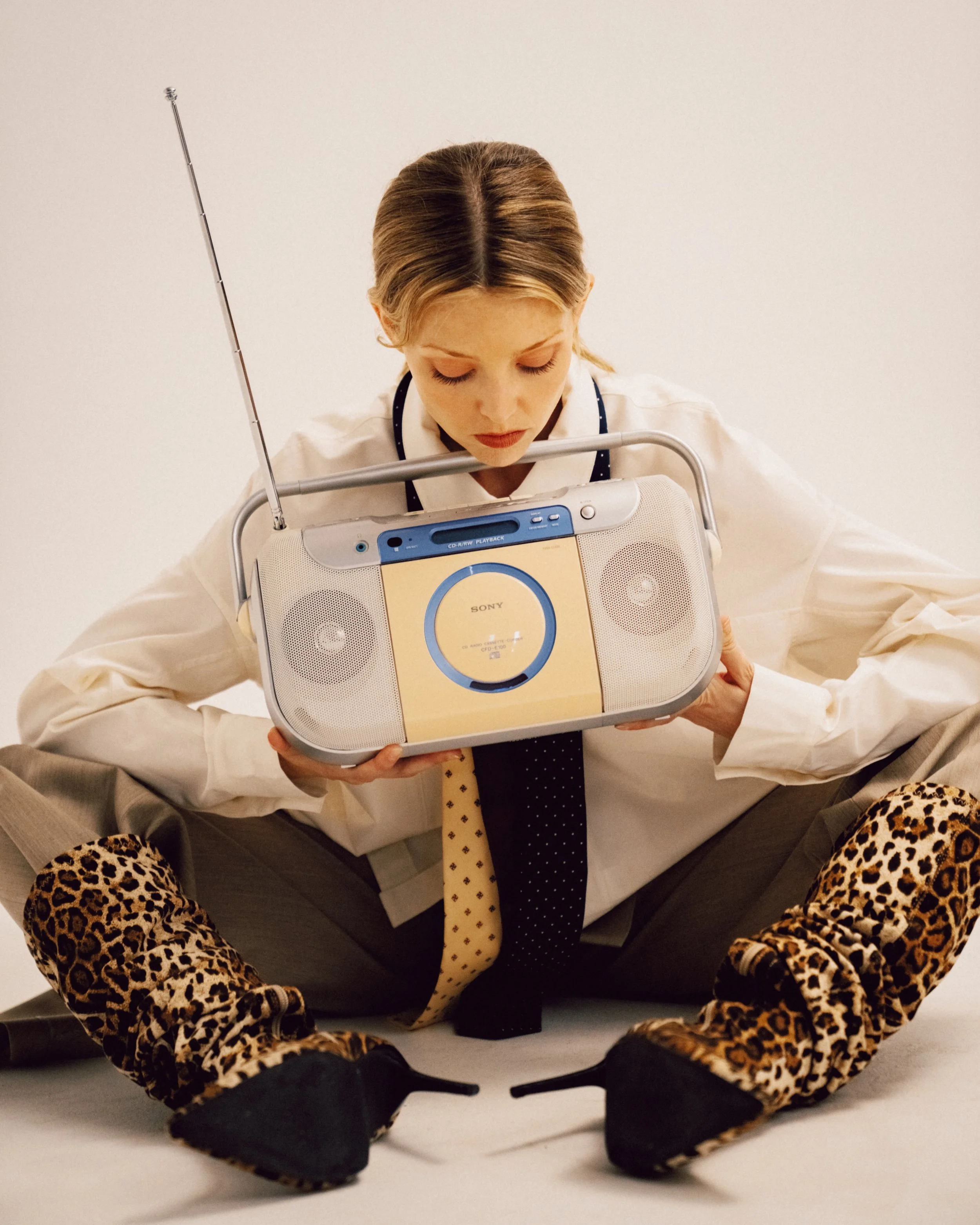 A blonde woman sitting on the floor holding a Sony boombox close to her face, wearing a white button-down shirt with ties, trousers, and leopard-print heeled boots.