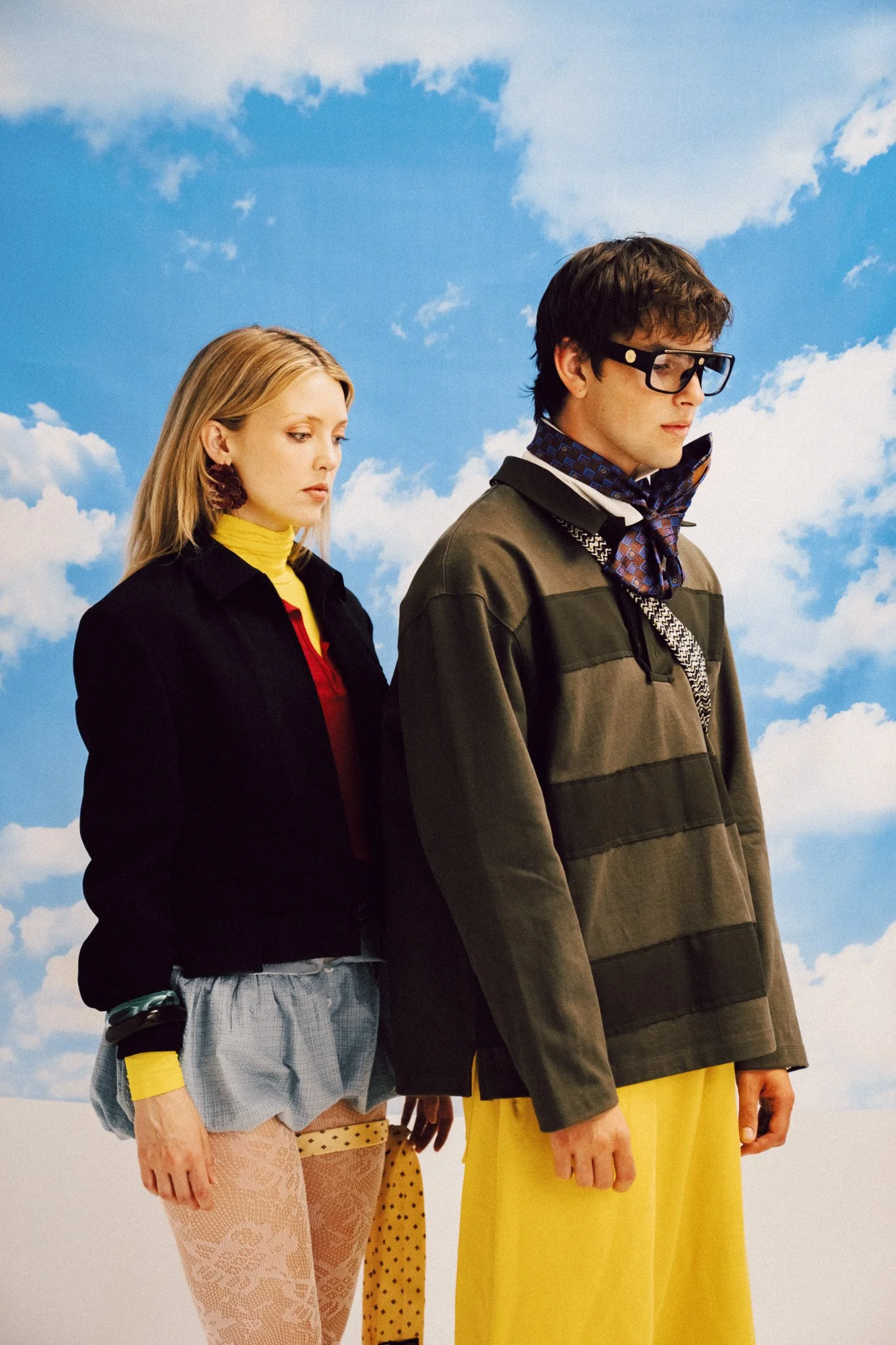 A portrait of a blonde woman and a young man standing side by side against a blue sky backdrop, both wearing layered outfits featuring colorful garments, patterns, and accessories.