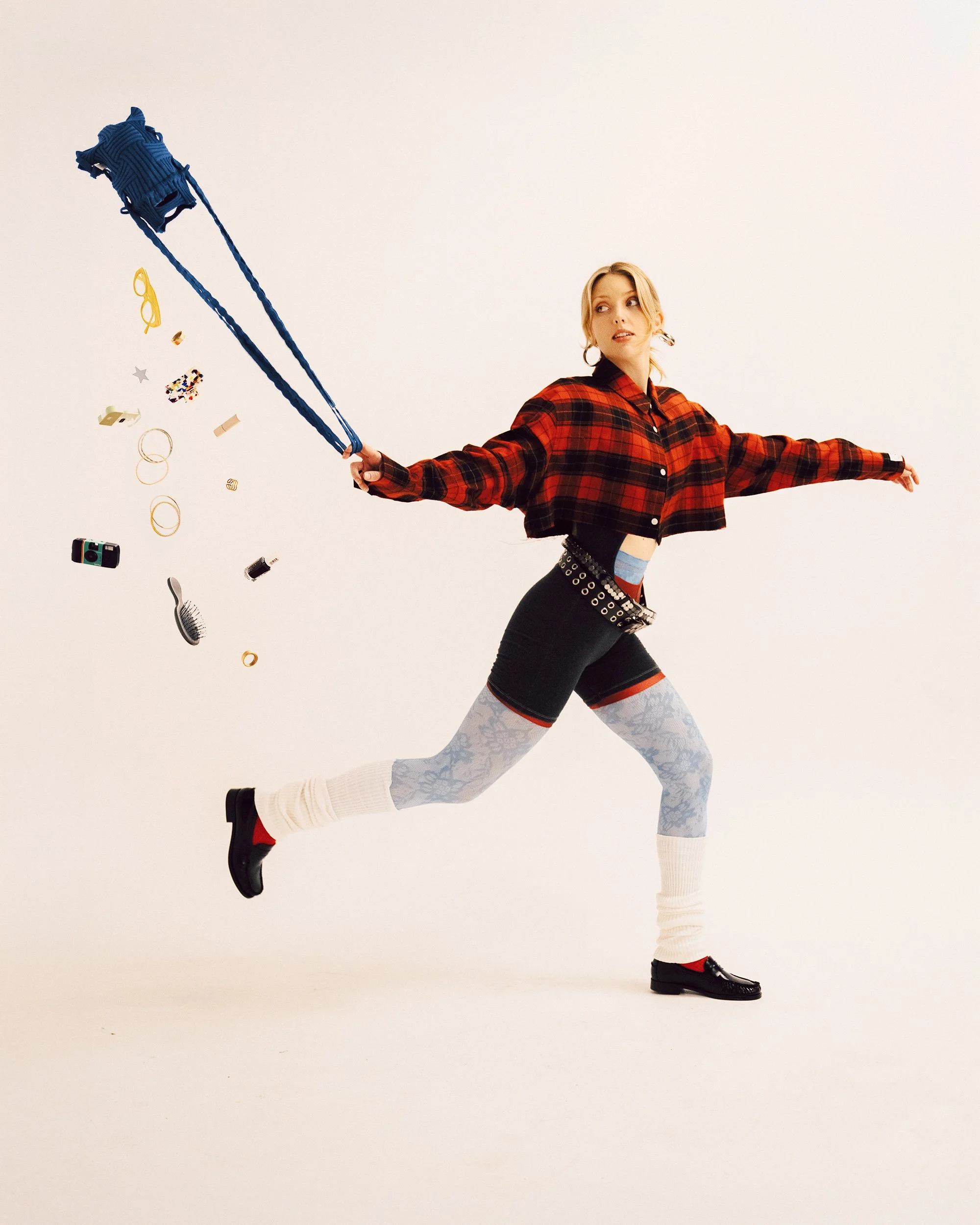 A blonde woman in a dynamic running pose looking back as small items tumble out of her bag, wearing a red and black plaid cropped button-down shirt, dark shorts over patterned tights, white leg warmers, loafers, double belts, and hoop earrings.