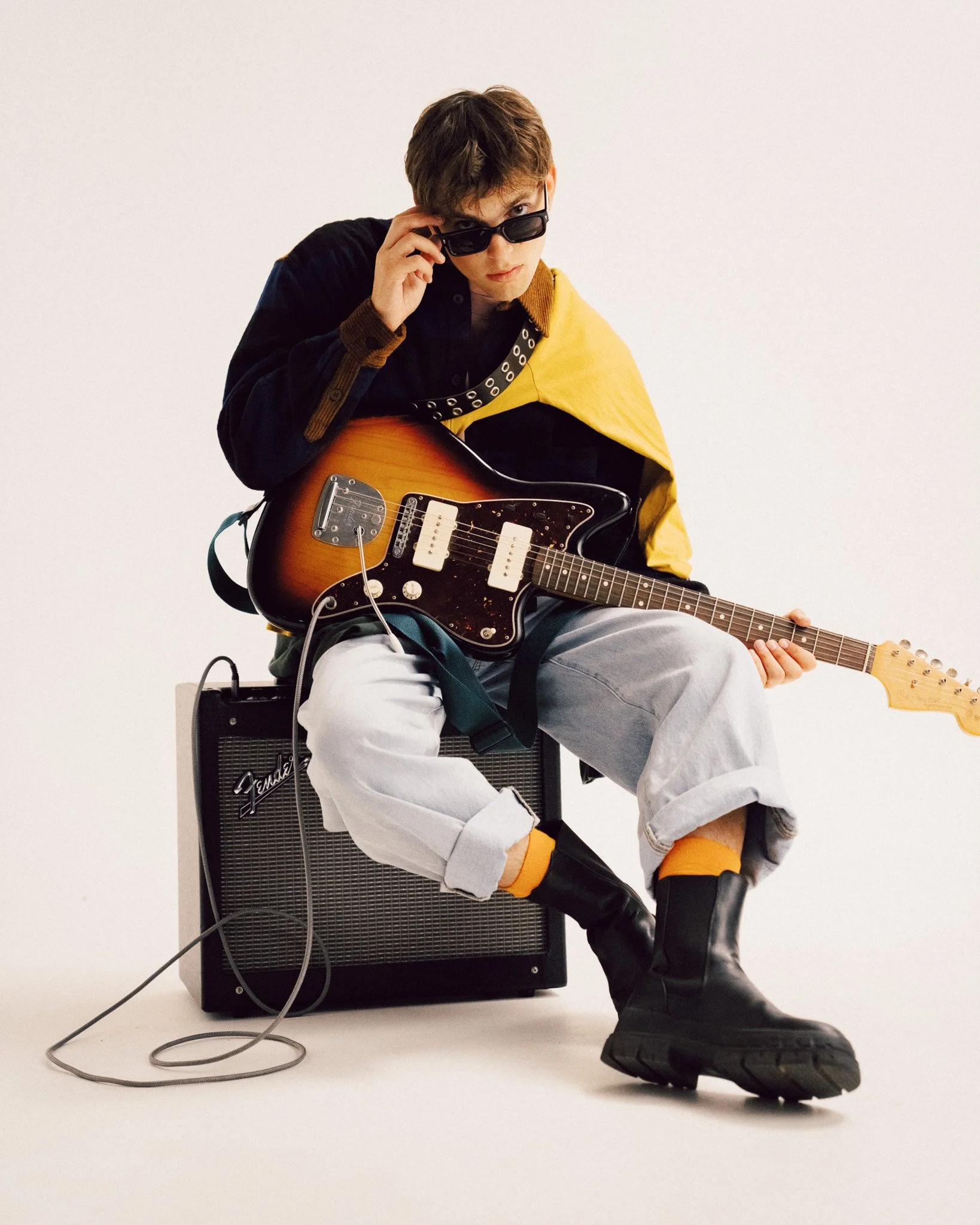 A young man sitting on an amplifier while holding an electric guitar and adjusting his sunglasses, wearing a dark patterned shirt with a contrasting collar, a yellow garment draped over his shoulder, light-colored cuffed jeans, socks, and dark boots.