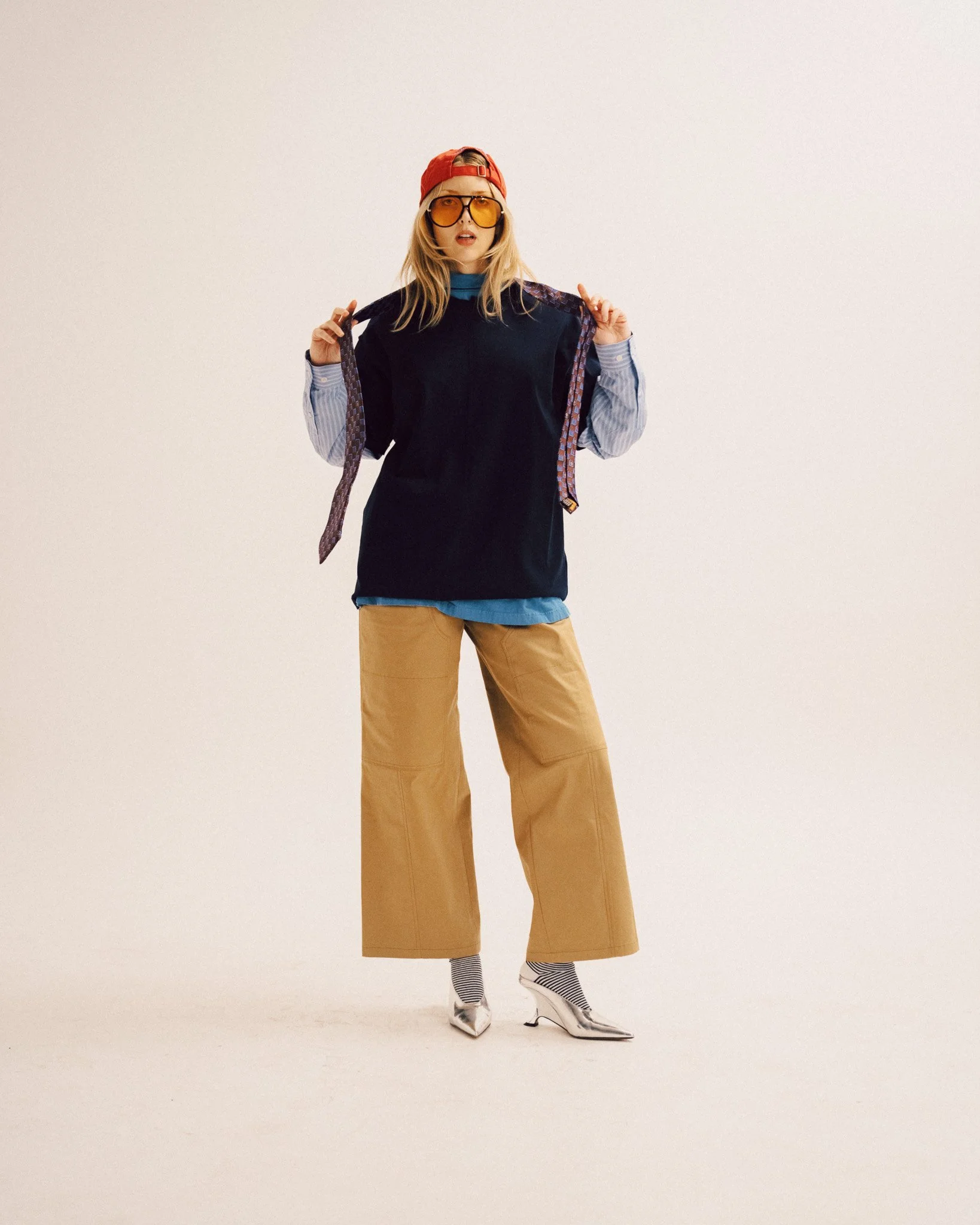 A blonde woman standing and holding a patterned necktie, wearing a dark top layered over a striped shirt, loose-fitting khaki trousers, striped socks, silver metallic shoes, tinted glasses, and a red cap.