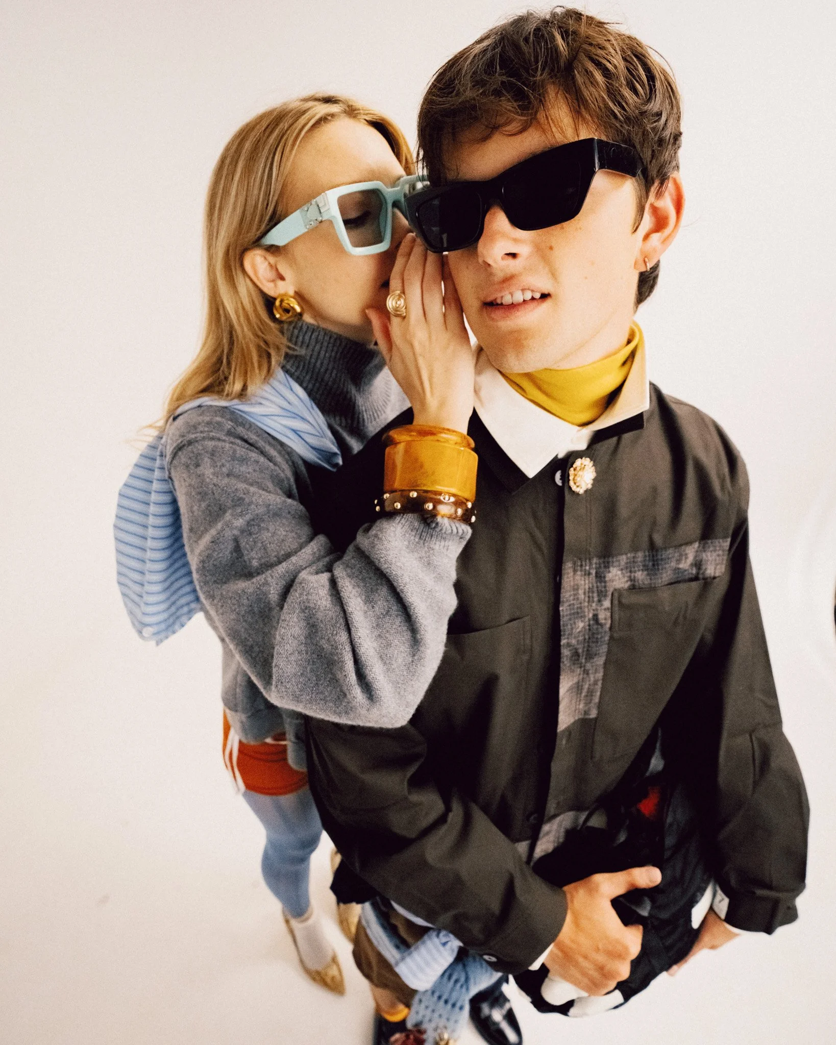 A close-up portrait of a blonde woman leaning in to whisper into the ear of a young man wearing sunglasses, both styled in layered outfits with accessories