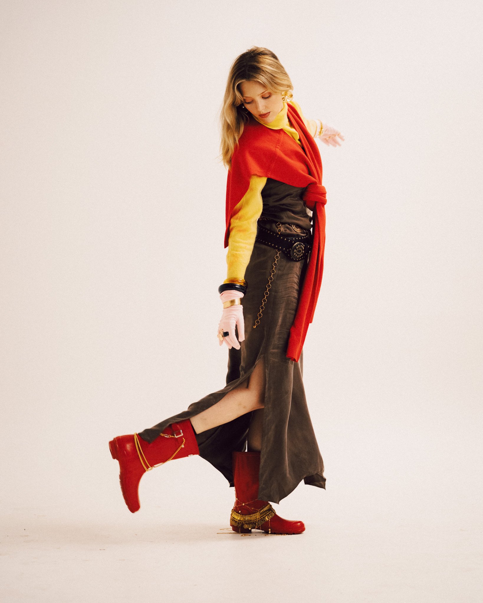A blonde woman posing dynamically with one leg lifted, wearing a dark skirt with a side slit, a layered yellow top with a red sweater draped over her shoulders, belts, accessories, and red boots.