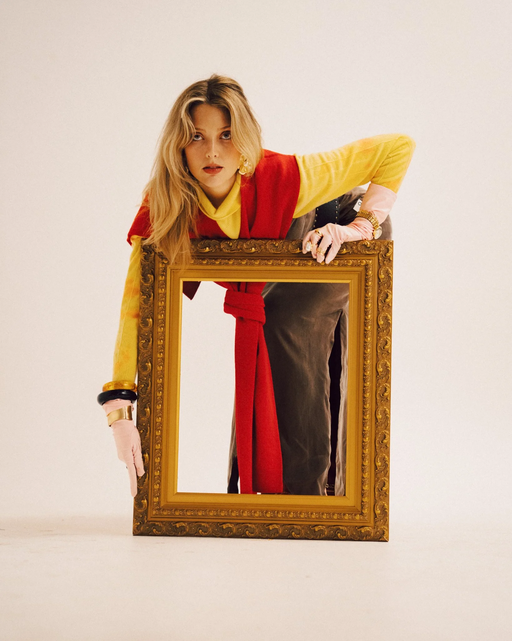 A blonde woman wearing a yellow turtleneck, red sweater layered over her shoulders, and pink gloves, leaning forward and posing behind an ornate antique gold picture frame.