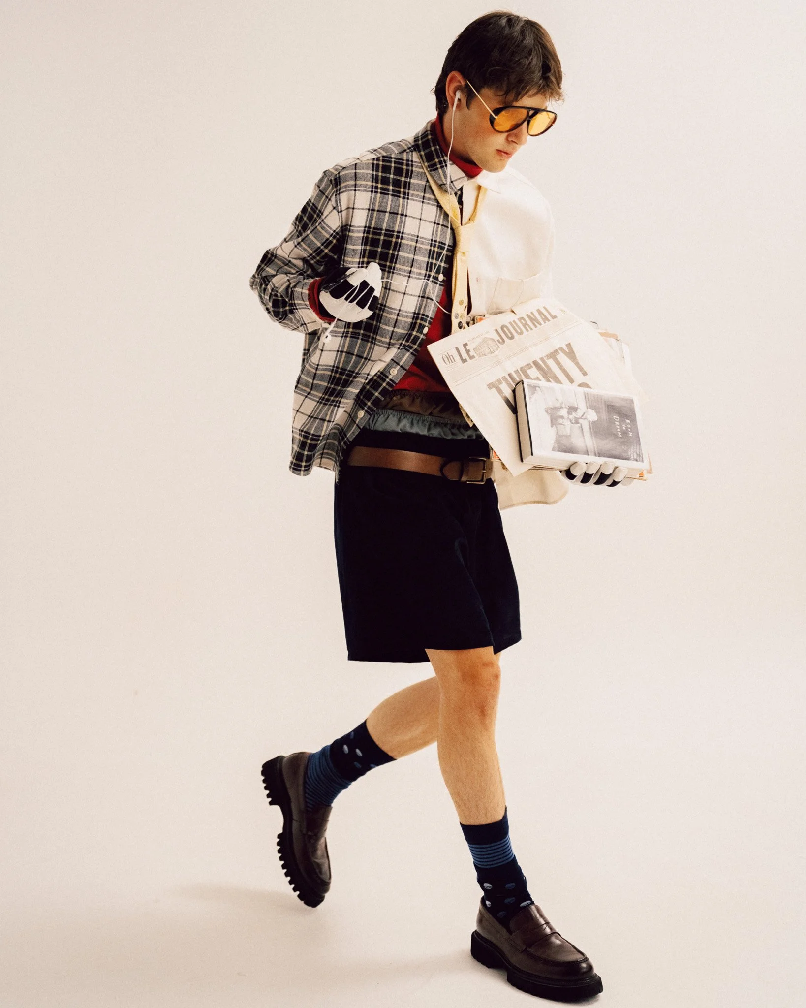A young man walking while carrying a publication and wearing a split-design plaid and white shirt, dark shorts, a belt, patterned socks, dark loafers, tinted glasses, and wired earphones.