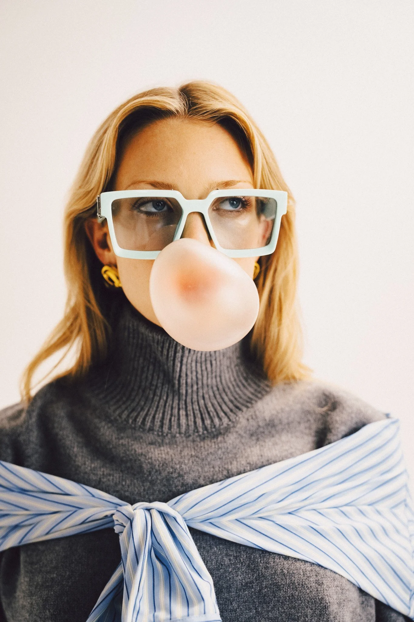 A close-up portrait of a blonde woman blowing a bubblegum bubble, wearing light-colored framed glasses, a grey turtleneck sweater, a striped garment tied around her shoulders, and statement earrings