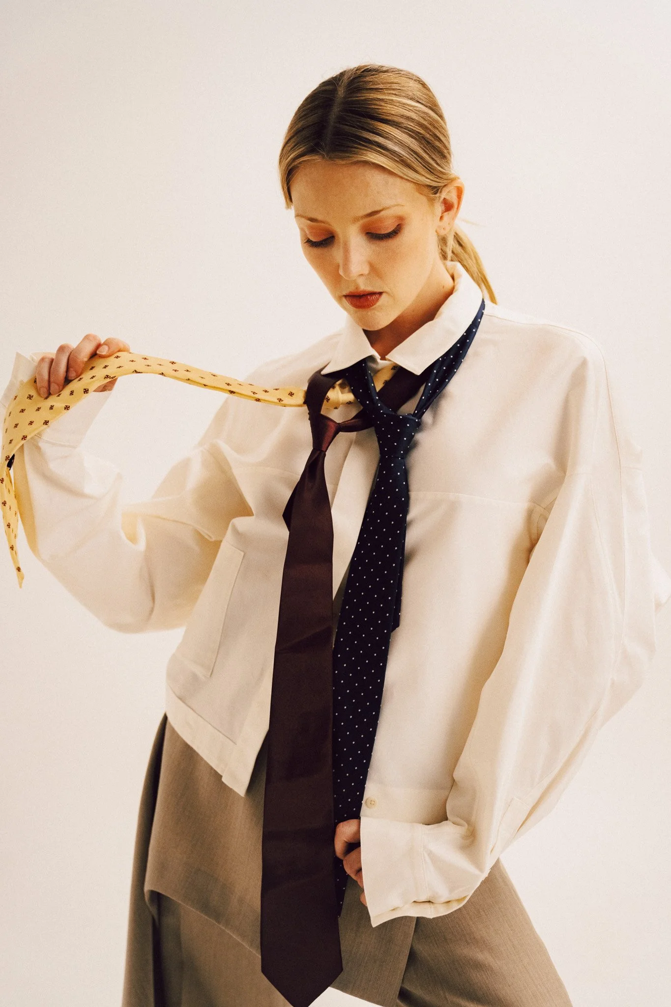 A blonde woman wearing an oversized white button-down shirt and grey trousers, looking down while holding and adjusting multiple ties draped around her neck.
