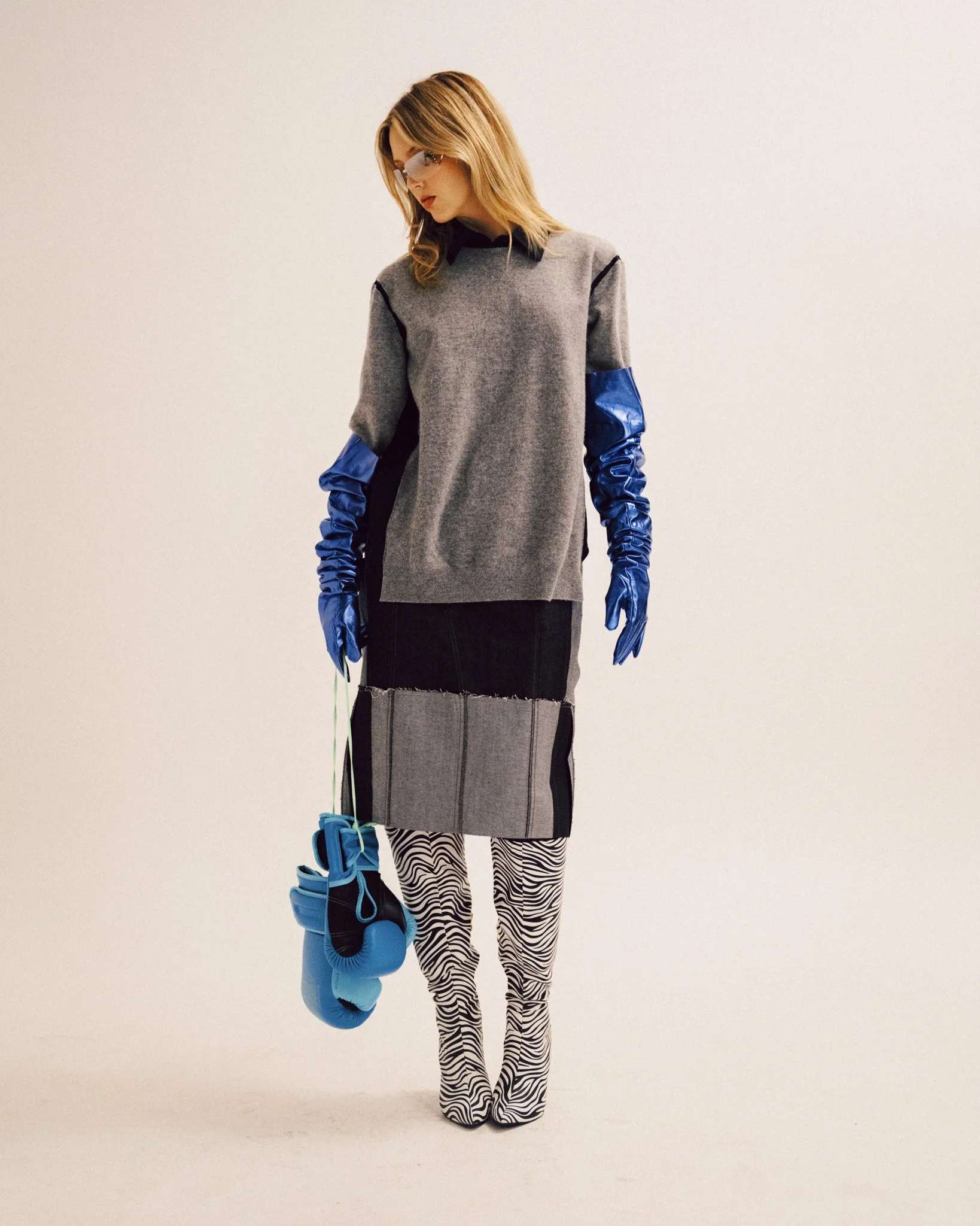 A blonde woman standing and looking down, wearing a grey knit top, a patchwork skirt, zebra print boots, shiny blue opera-length gloves, and holding a pair of blue boxing gloves.