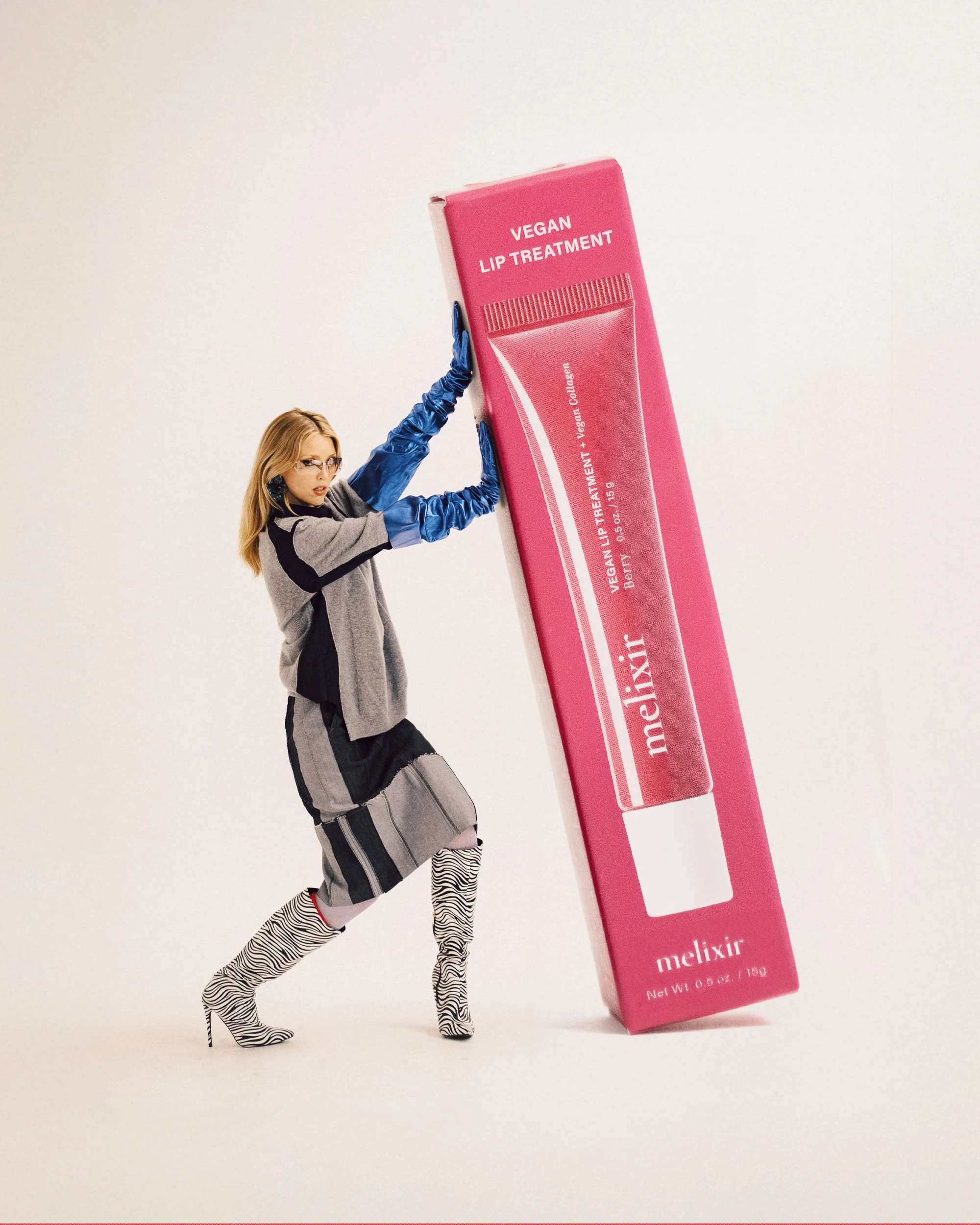 A blonde woman leaning against and holding up a giant pink 'Melixir Vegan Lip Treatment' product box, wearing a grey and dark outfit with blue gloves, glasses, and zebra-print boots.
